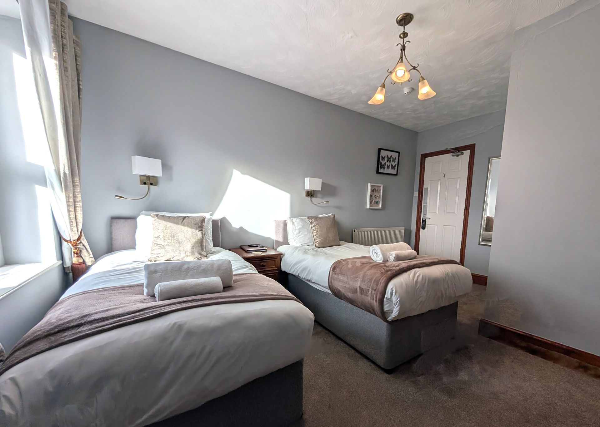 Twin-bedded hotel room with gray walls, two beds with white linens, window with curtains, and a door.