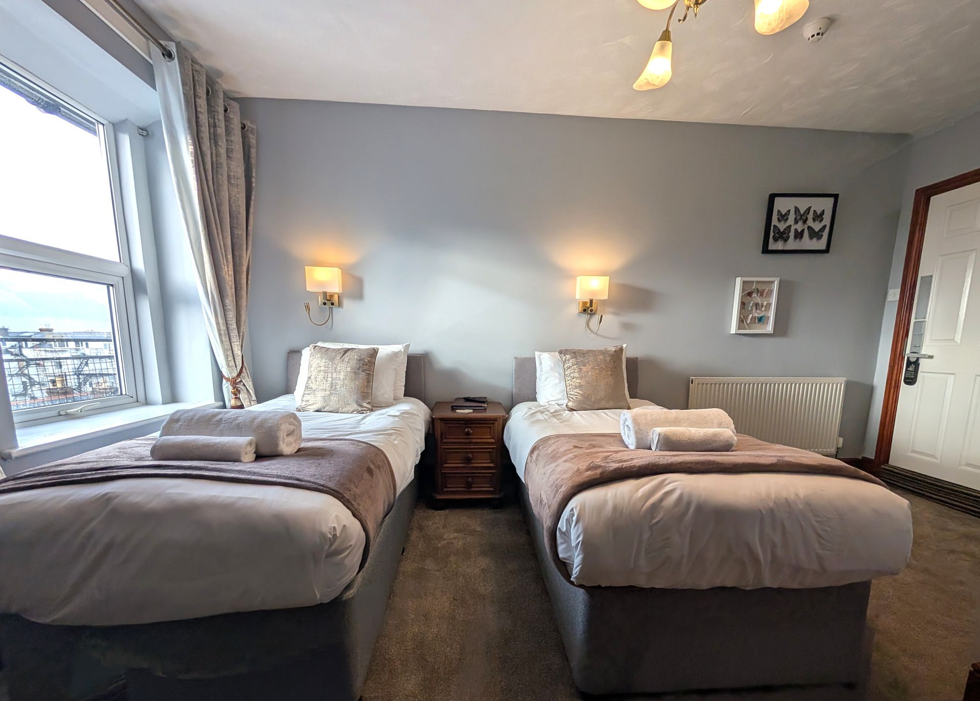 Two twin beds with beige and white bedding in a room with a window, lamps, and a door.