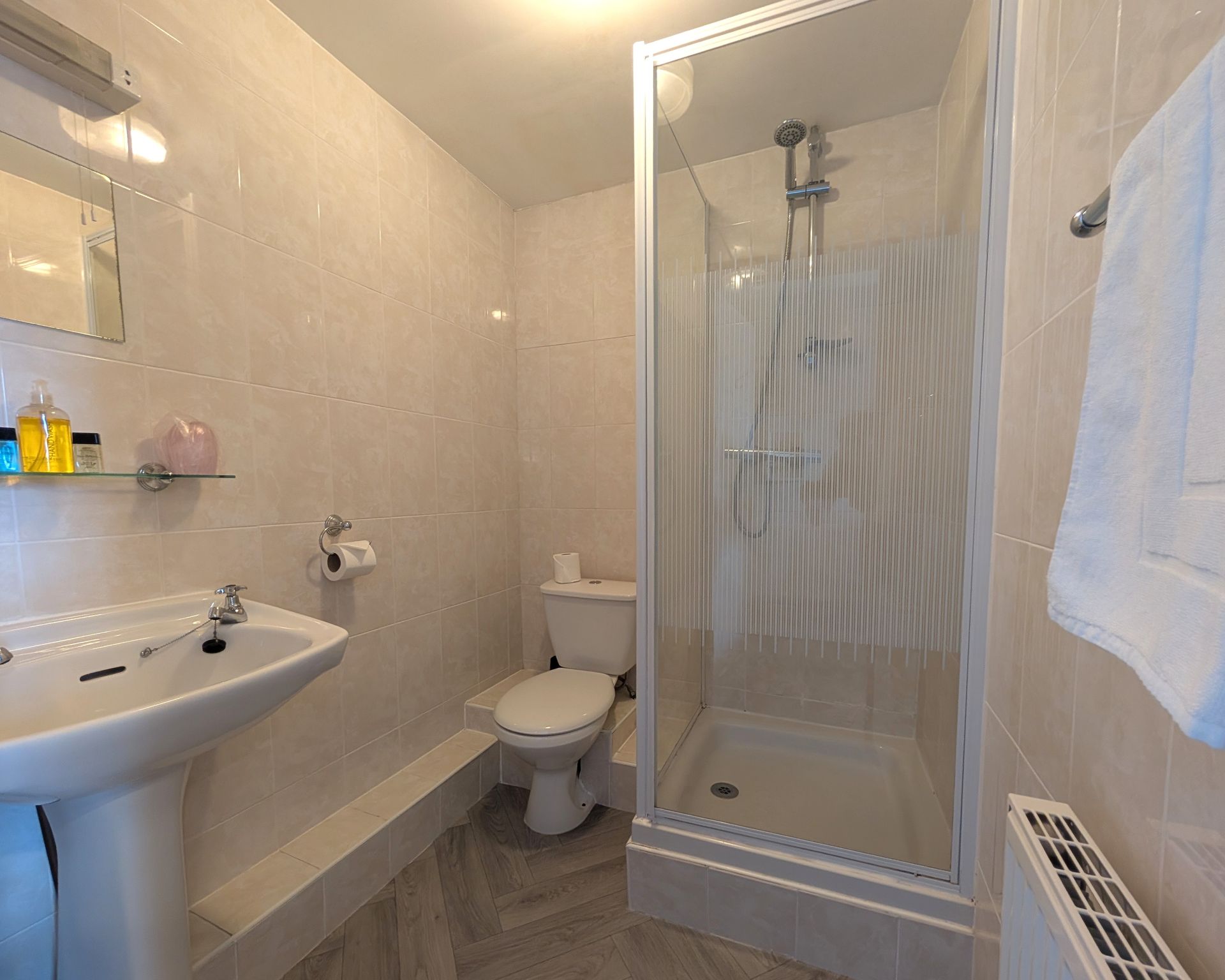 Bathroom with a pedestal sink, toilet, shower, and a towel on a radiator. Light-colored tiles cover the walls.