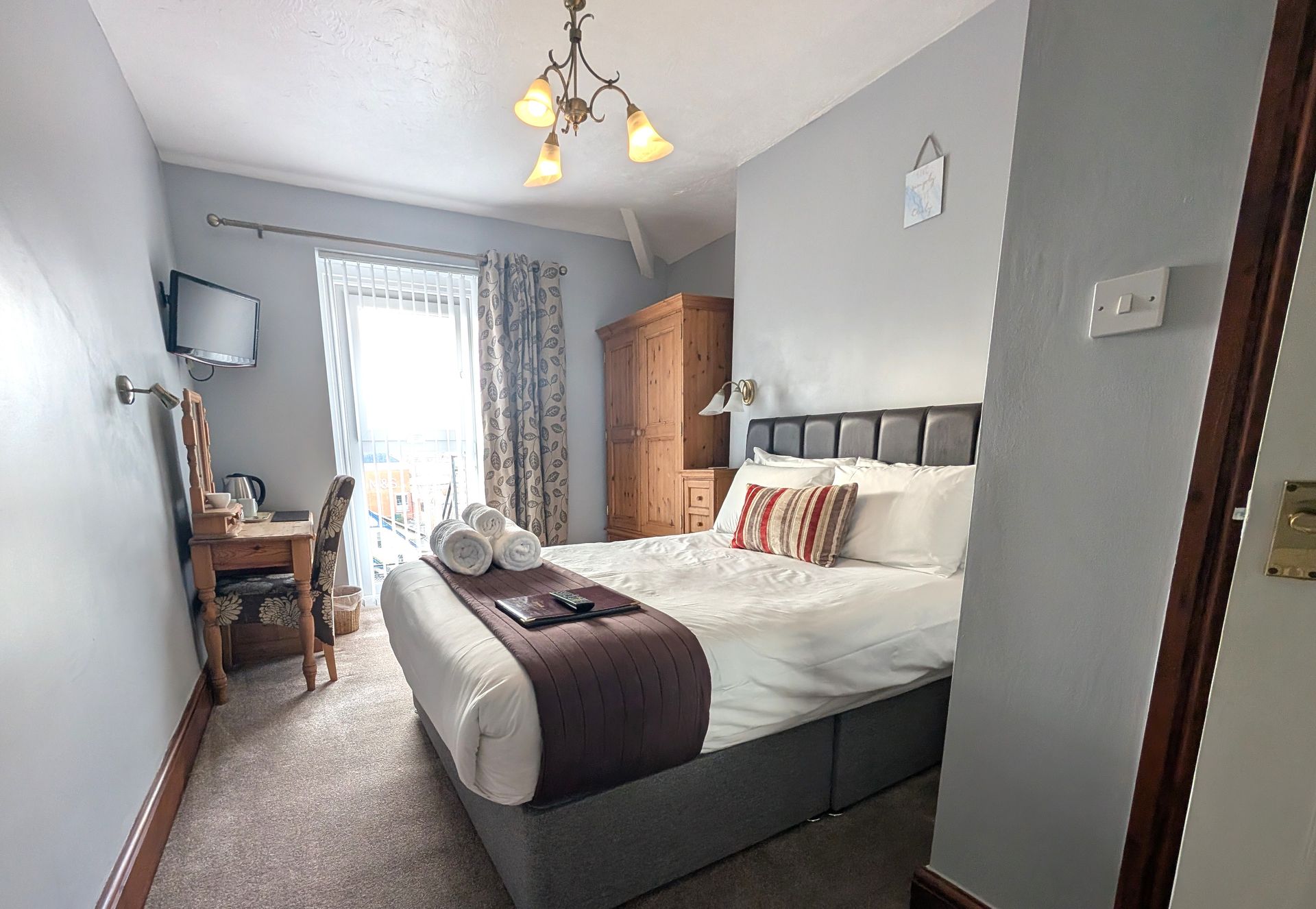 Bedroom with a double bed, wardrobe, desk, and balcony. Soft colors with light streaming in.