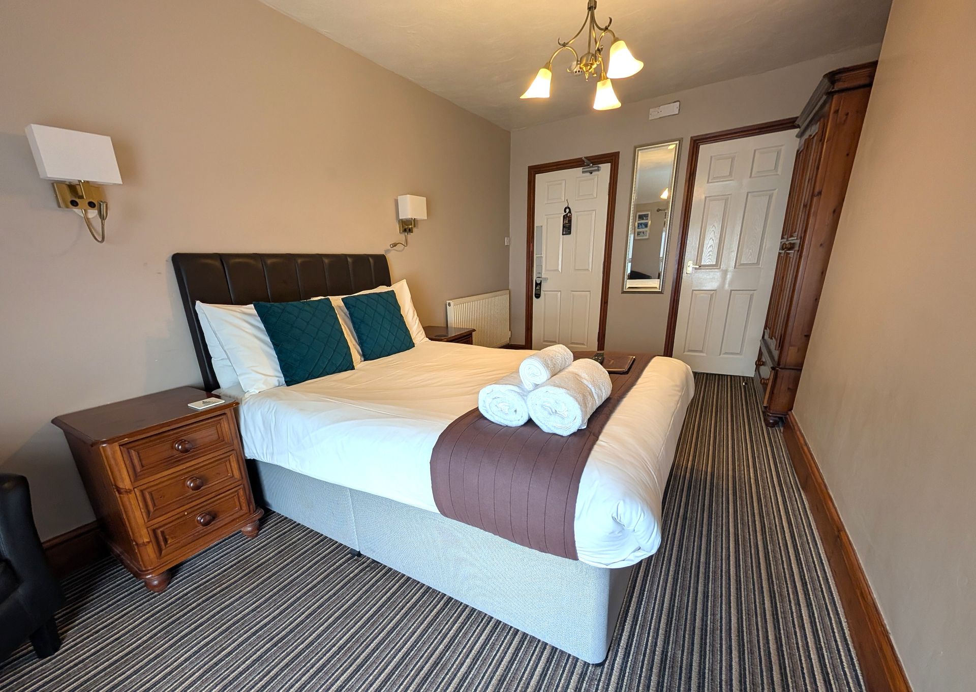 Bedroom with a bed, nightstand, and wardrobe. Beige walls, brown and white bedspread, two green pillows, three white towels.
