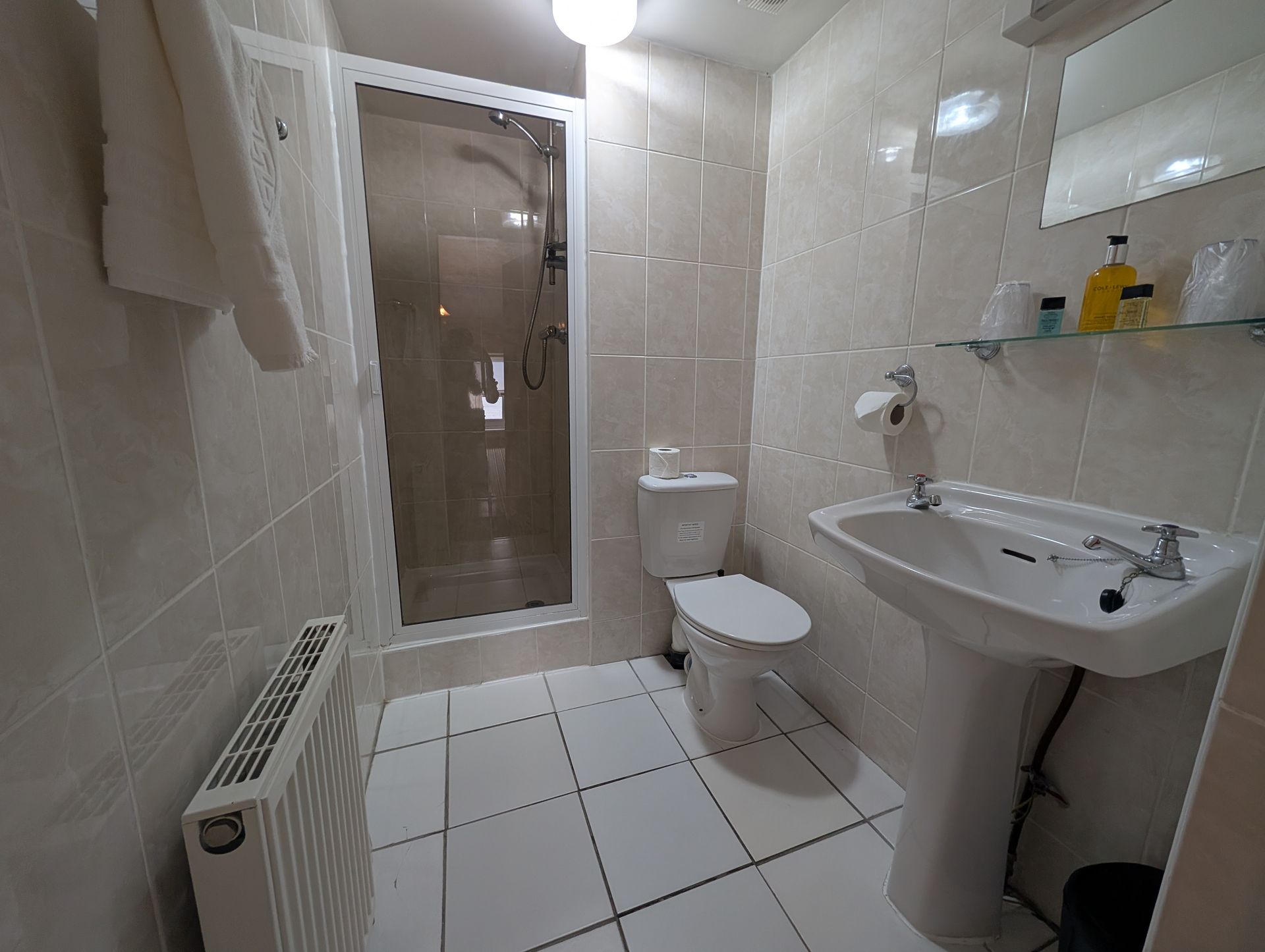 Small bathroom with white tiled walls and floor, shower, toilet, pedestal sink, radiator, and shelf.