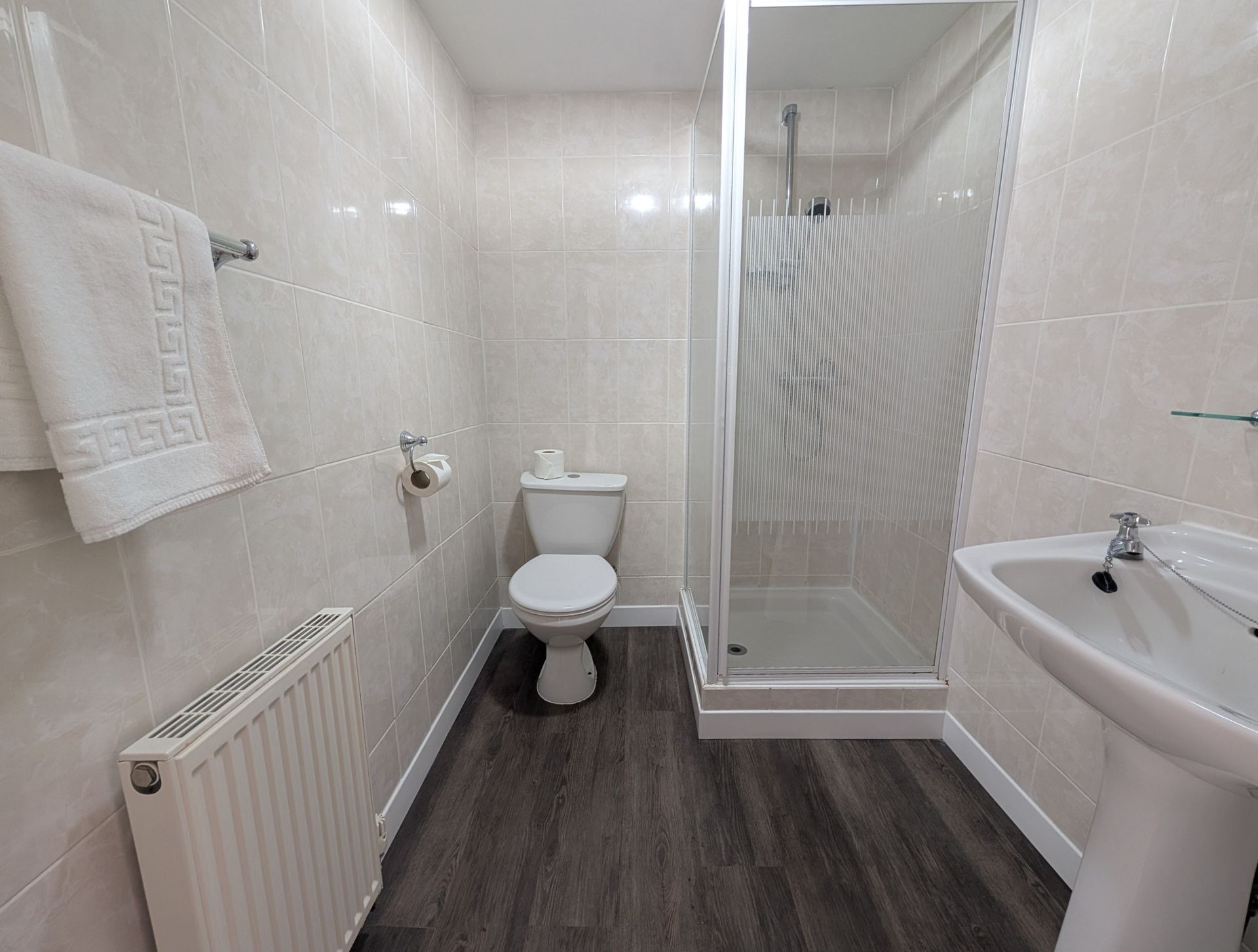 Bathroom with white tile walls, a toilet, shower, sink, radiator, and a towel on a rack.