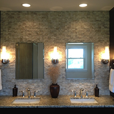 Bathroom with two sinks, mirrors, sconce lighting on stone textured wall, dark countertop.