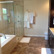 Bathroom with a glass shower, bathtub, and a towel rack with white robes. Brown tile floor.