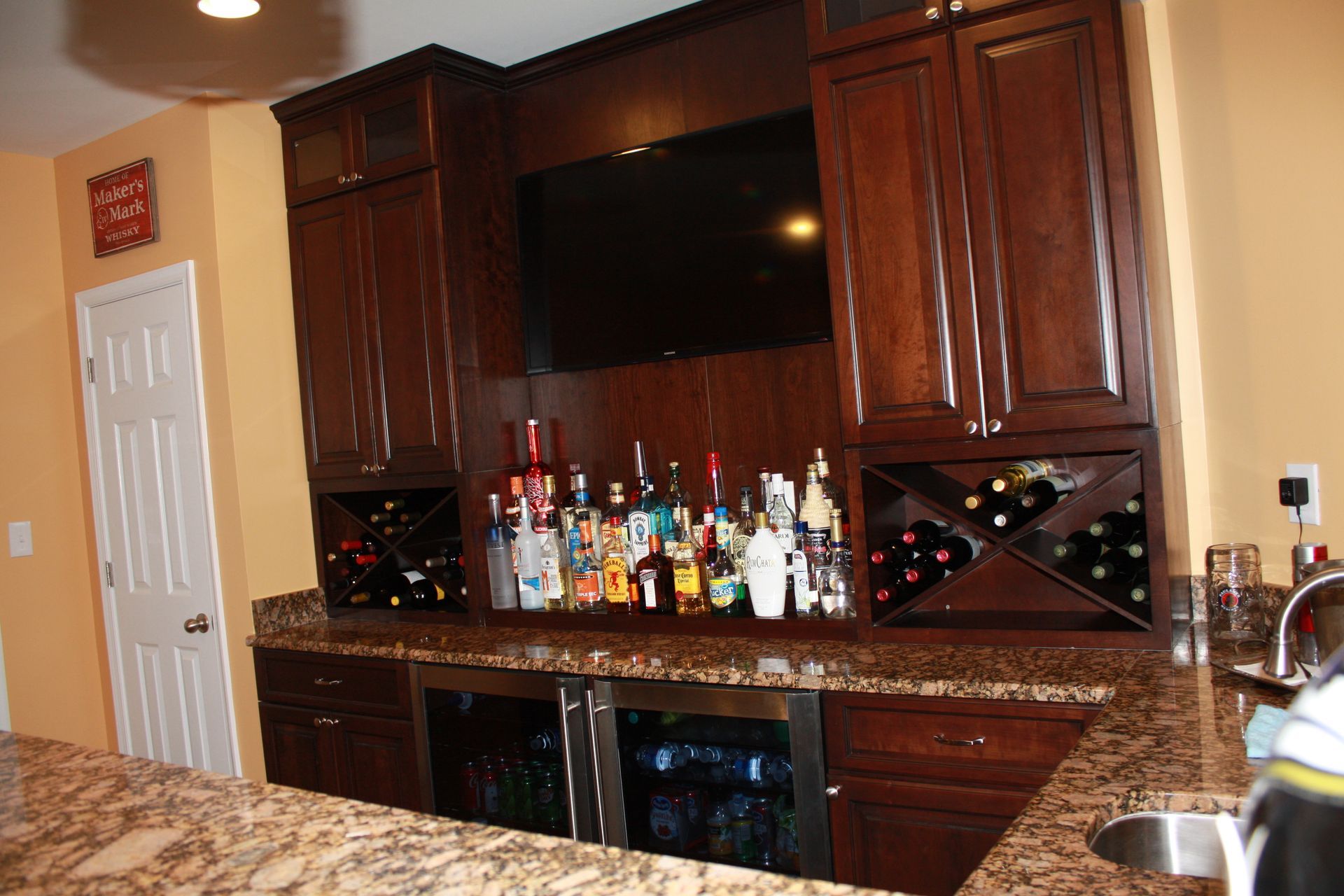 Built-in home bar with dark wood cabinets, granite countertops, wine racks, and a mounted TV, against tan walls.