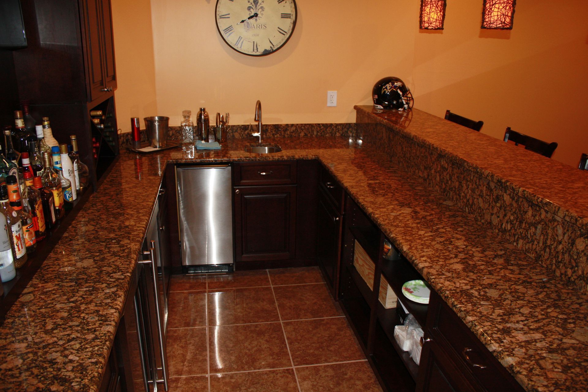 A home bar with dark wood cabinets and granite countertops, stainless steel appliances, and a large clock on the wall.