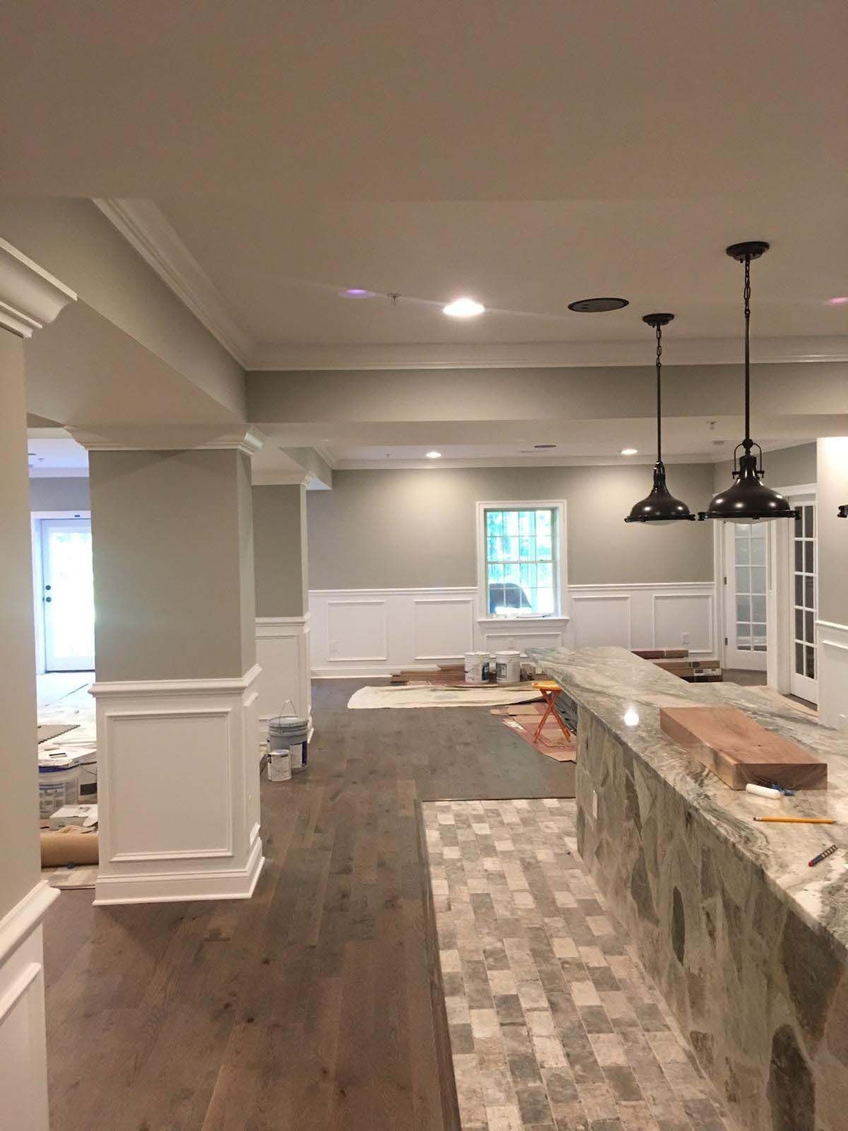Interior view of a renovated basement with a bar, dark pendant lights, gray walls, white wainscoting and dark wood floors.