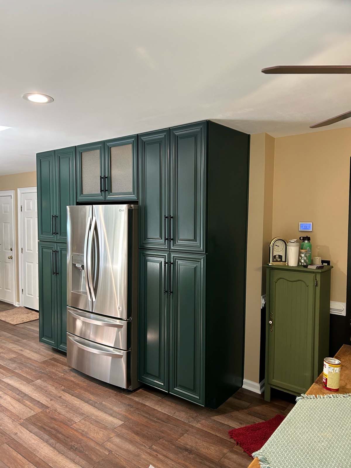 Green cabinets surround a stainless steel refrigerator in a kitchen with wood floors.