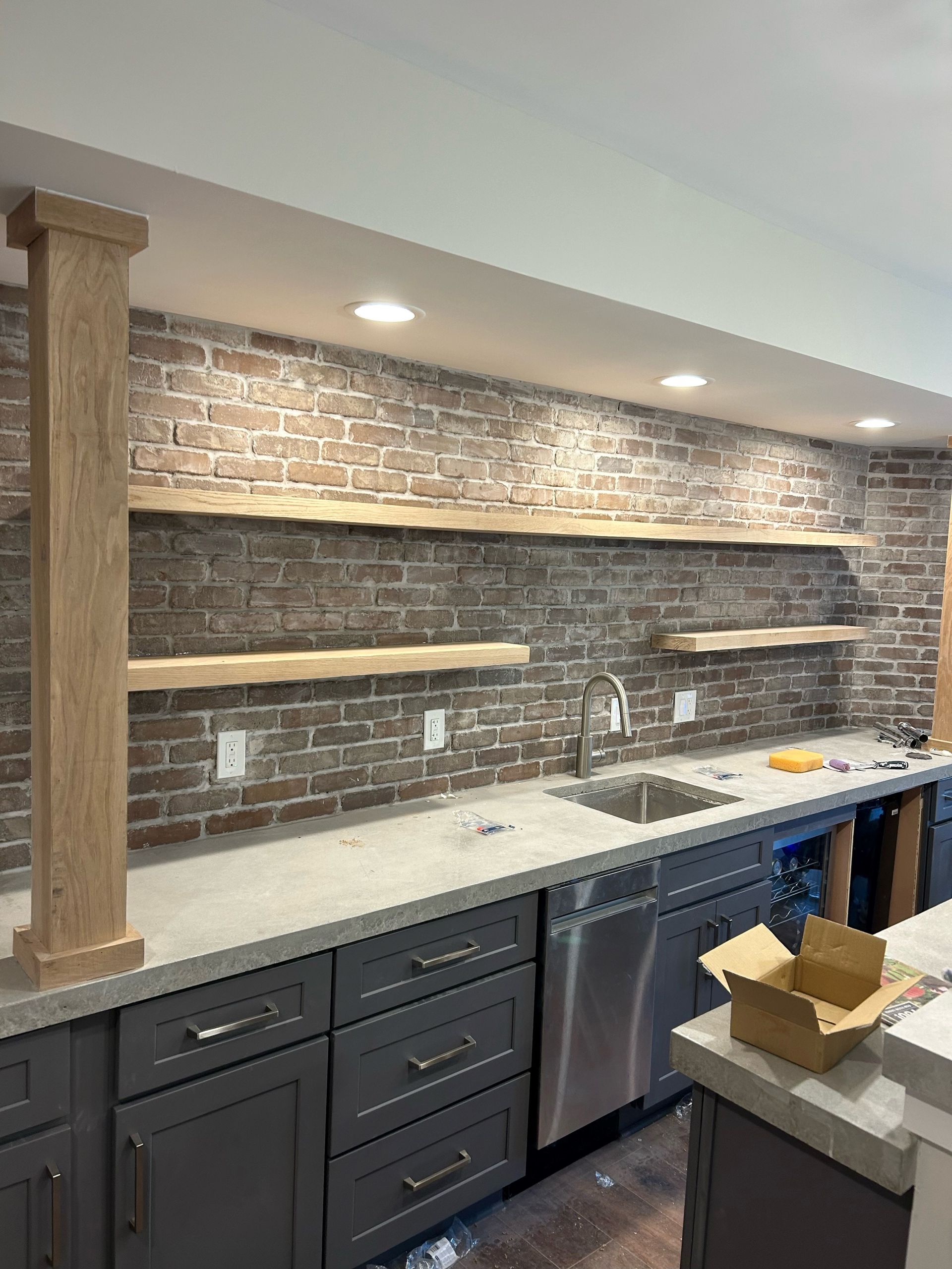 Gray kitchen with exposed brick wall, floating shelves, cabinets, sink, and stainless steel appliances.