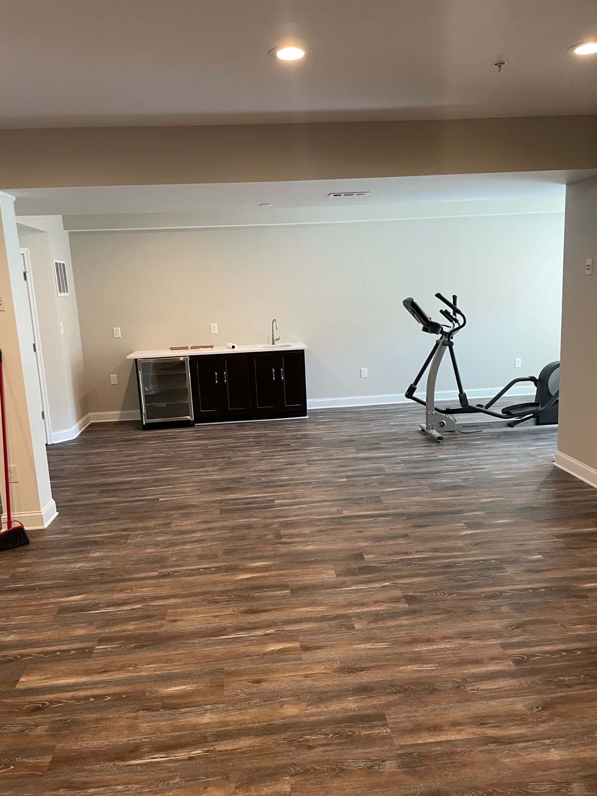 A mostly empty room with wood-look flooring, an elliptical machine, and a dark-colored cabinet with a refrigerator.