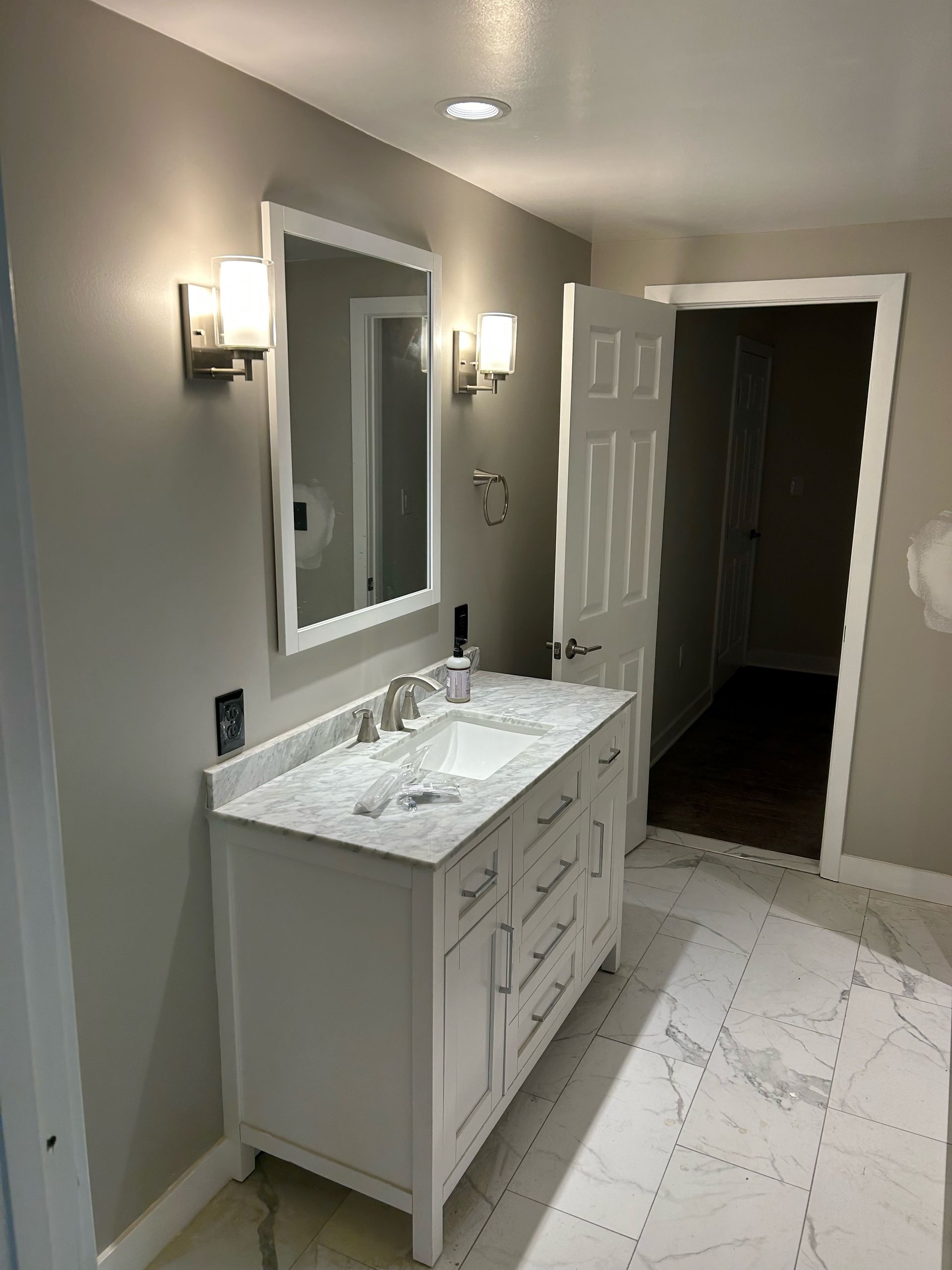 A renovated bathroom with a white vanity, mirror, and marble-like flooring; light gray walls and a doorway.