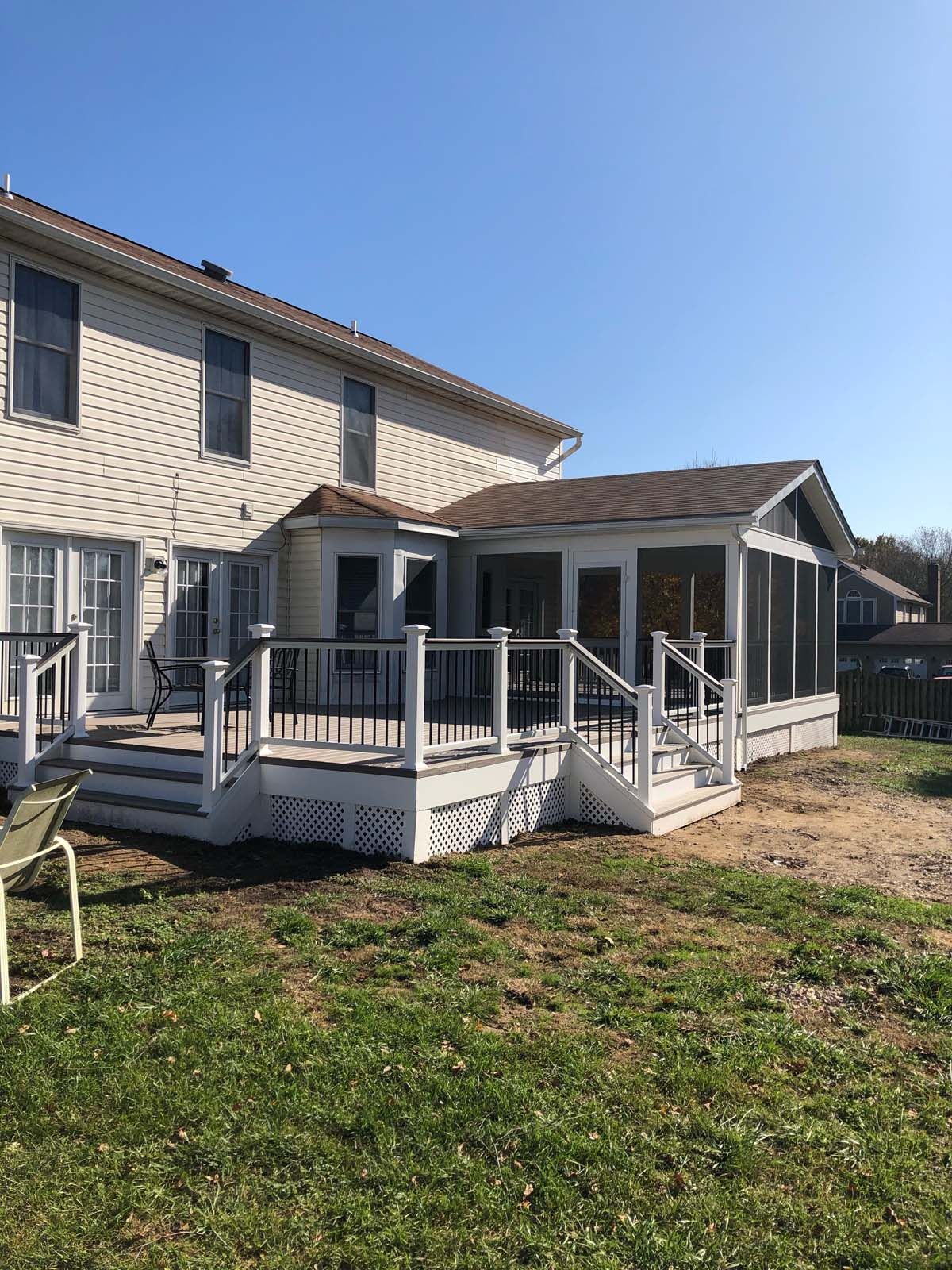 A two-story house with a white deck and screened-in porch. Green grass in the foreground, clear blue sky.