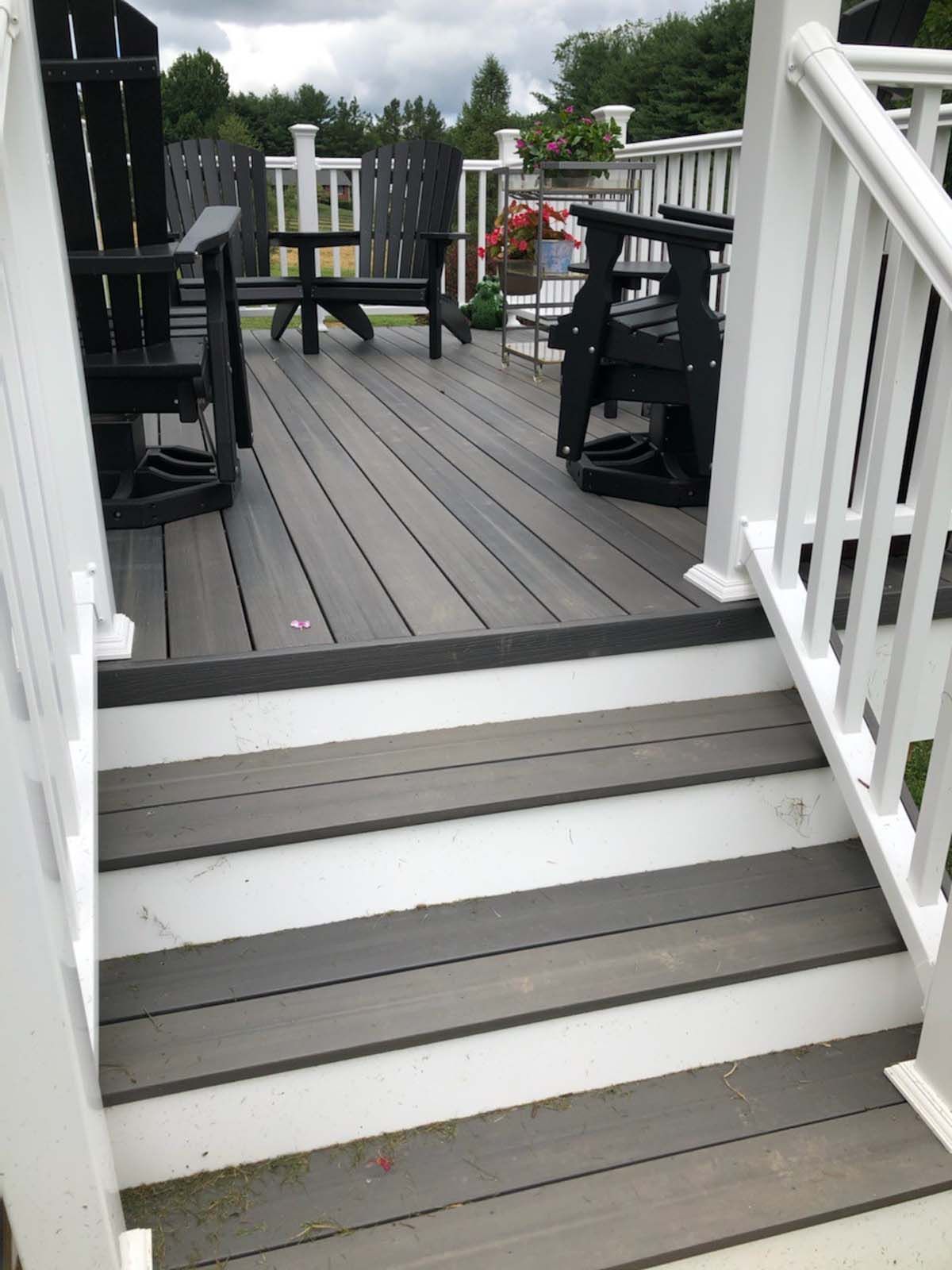 A deck with white railings, gray steps and wooden planks, and black Adirondack chairs.