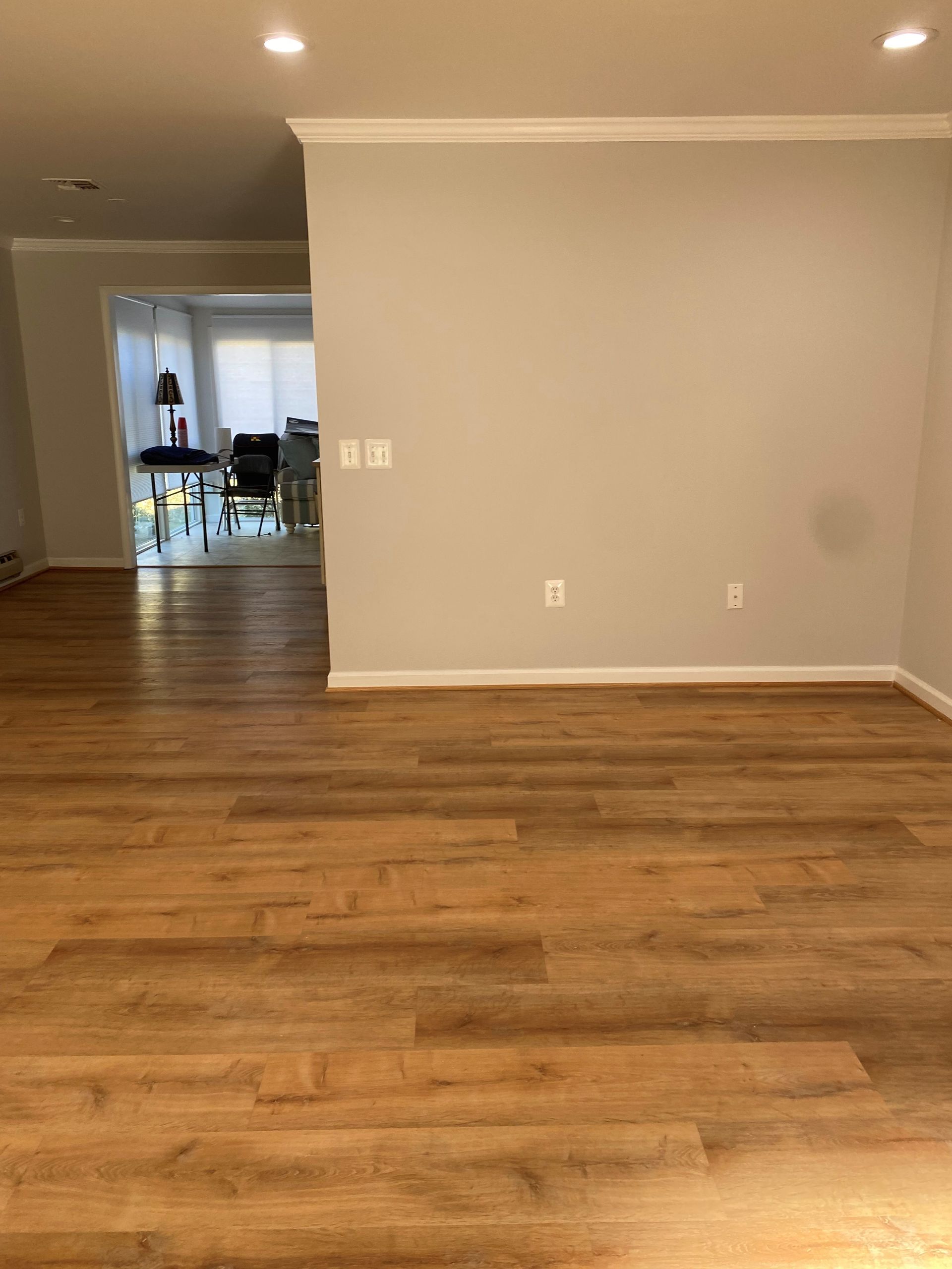 Hardwood floors in a living room with light gray walls and a view into another room with a desk and chairs.