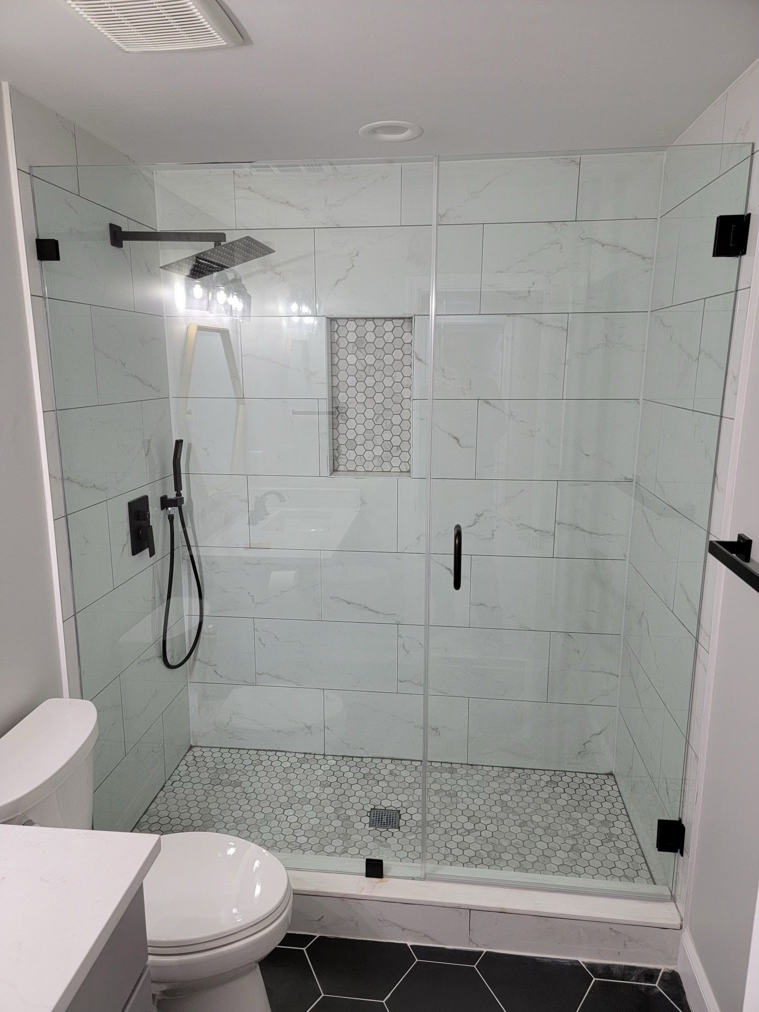 Modern white tiled shower with glass doors, black hardware, and a mosaic accent. Toilet and vanity.