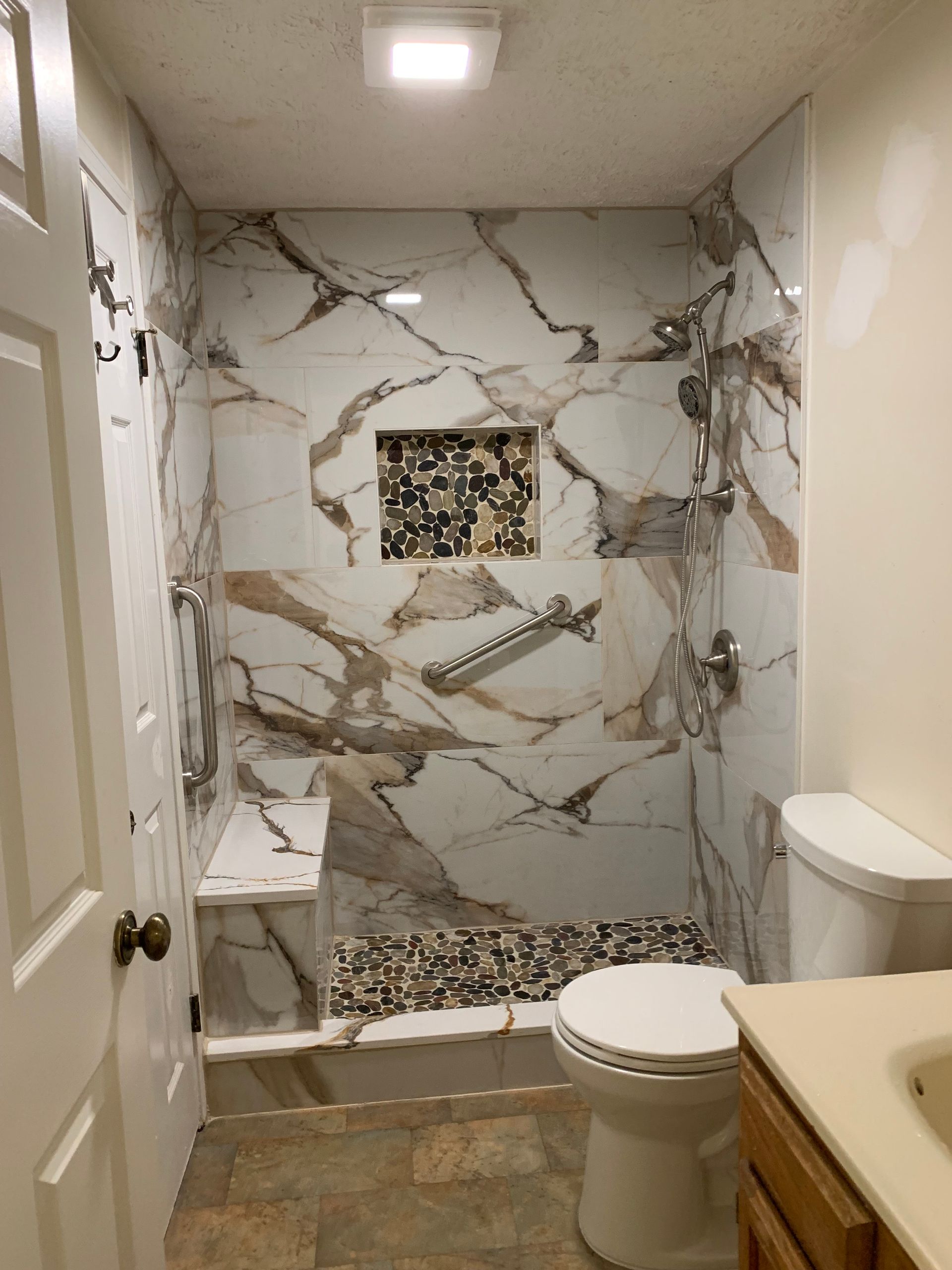 Bathroom with marble-like tiled shower, built-in seat, pebble floor, and toilet. Neutral walls.
