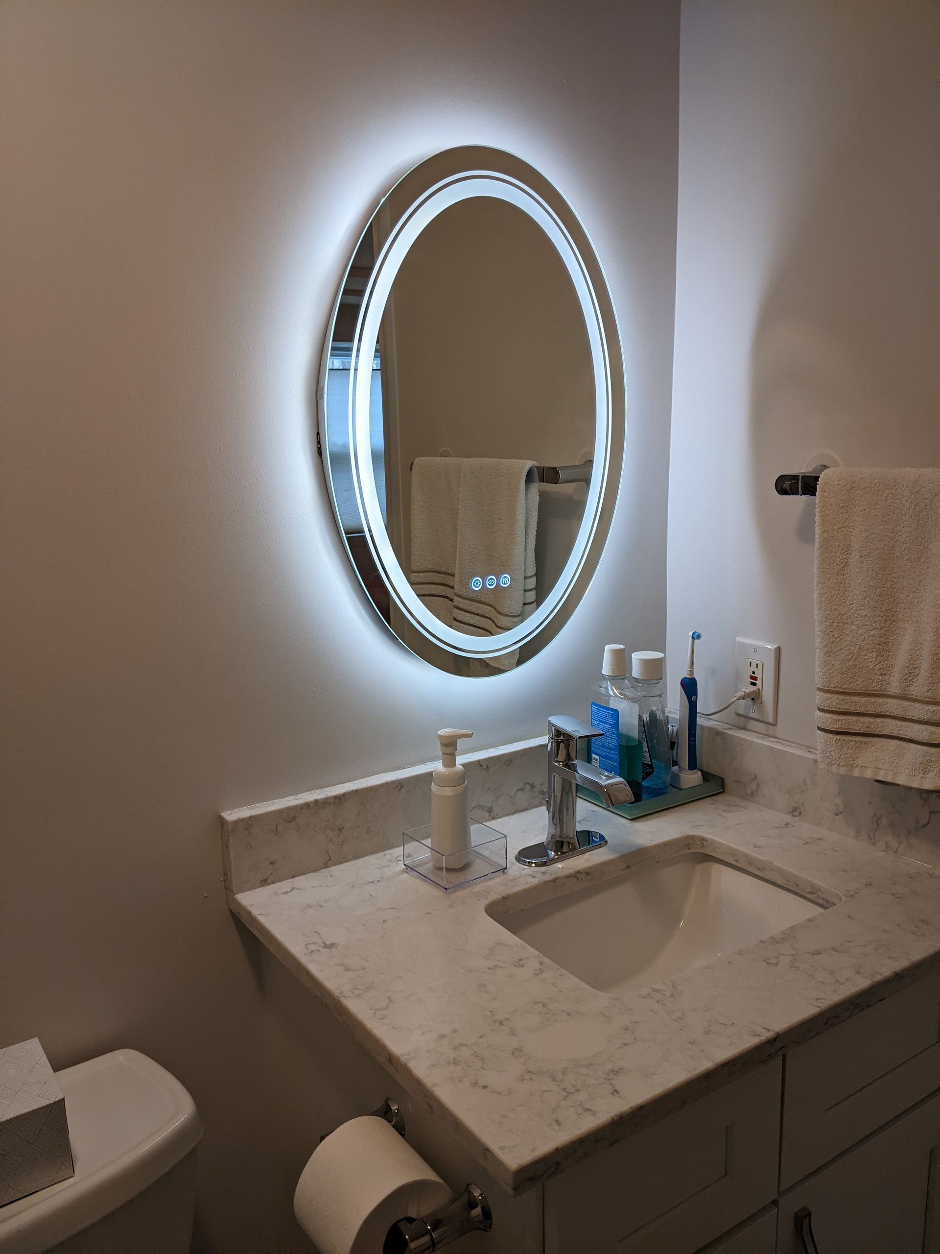 Bathroom with oval, illuminated mirror above a sink and countertop. A towel rack and toilet are visible.