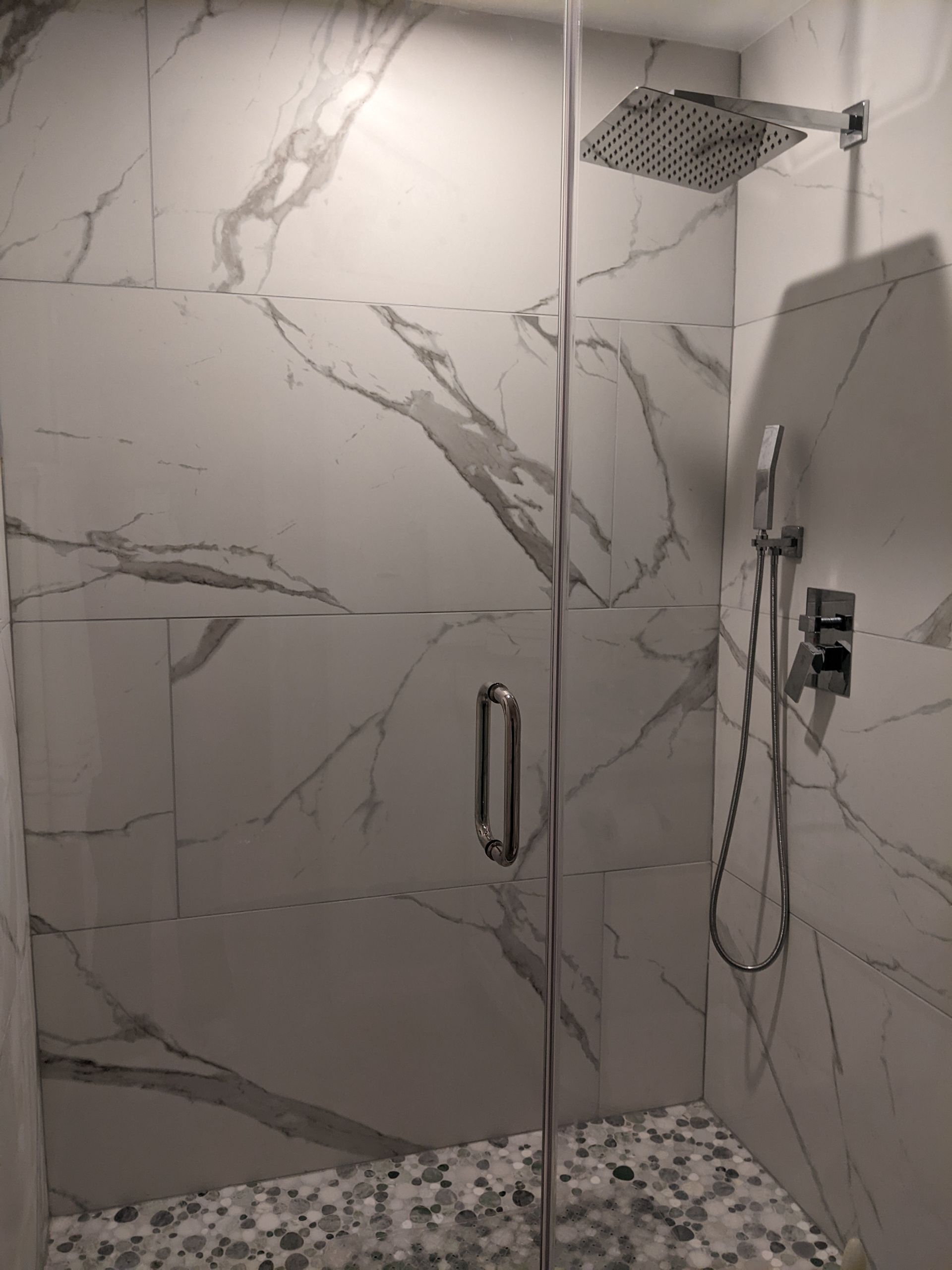 Shower with glass door, marble-look tile walls and mosaic floor. Silver showerhead and handheld sprayer.