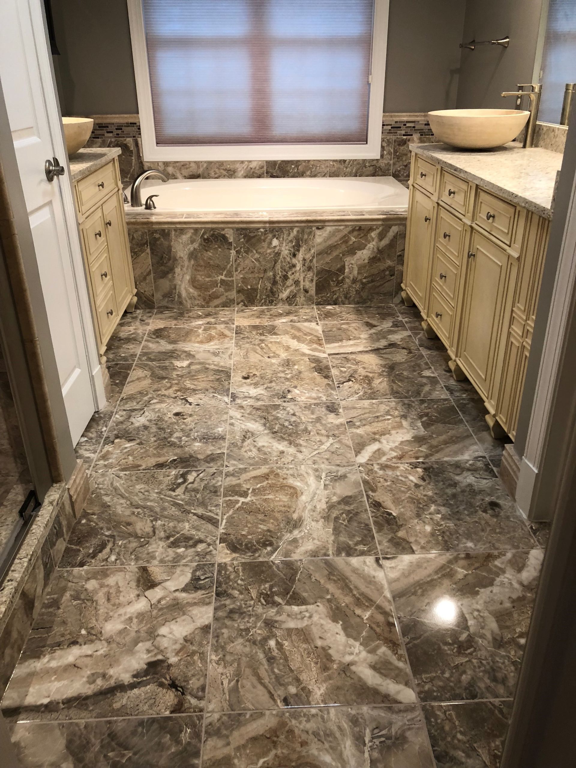 Bathroom with marble-like tile floor, tub, cream-colored cabinets, and a window with blinds.