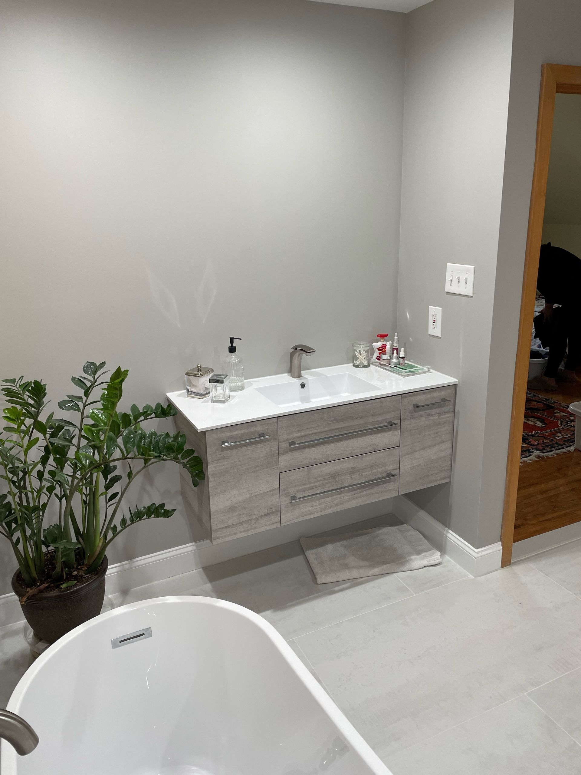 Bathroom with floating vanity, bathtub, and plant. Light gray walls, light wood vanity, and white tub.