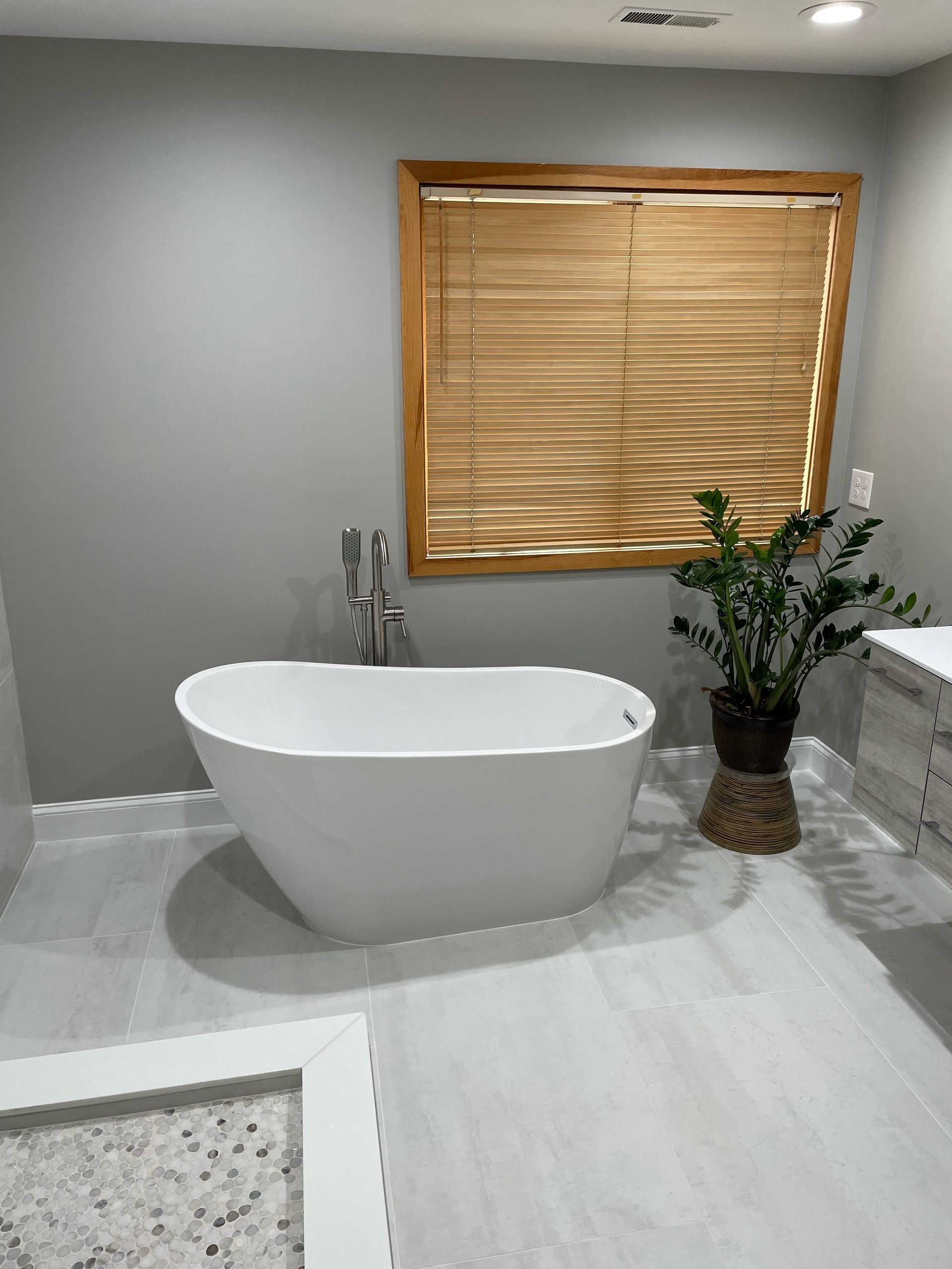 Modern bathroom with white tub, gray walls, wood blinds, and plant.