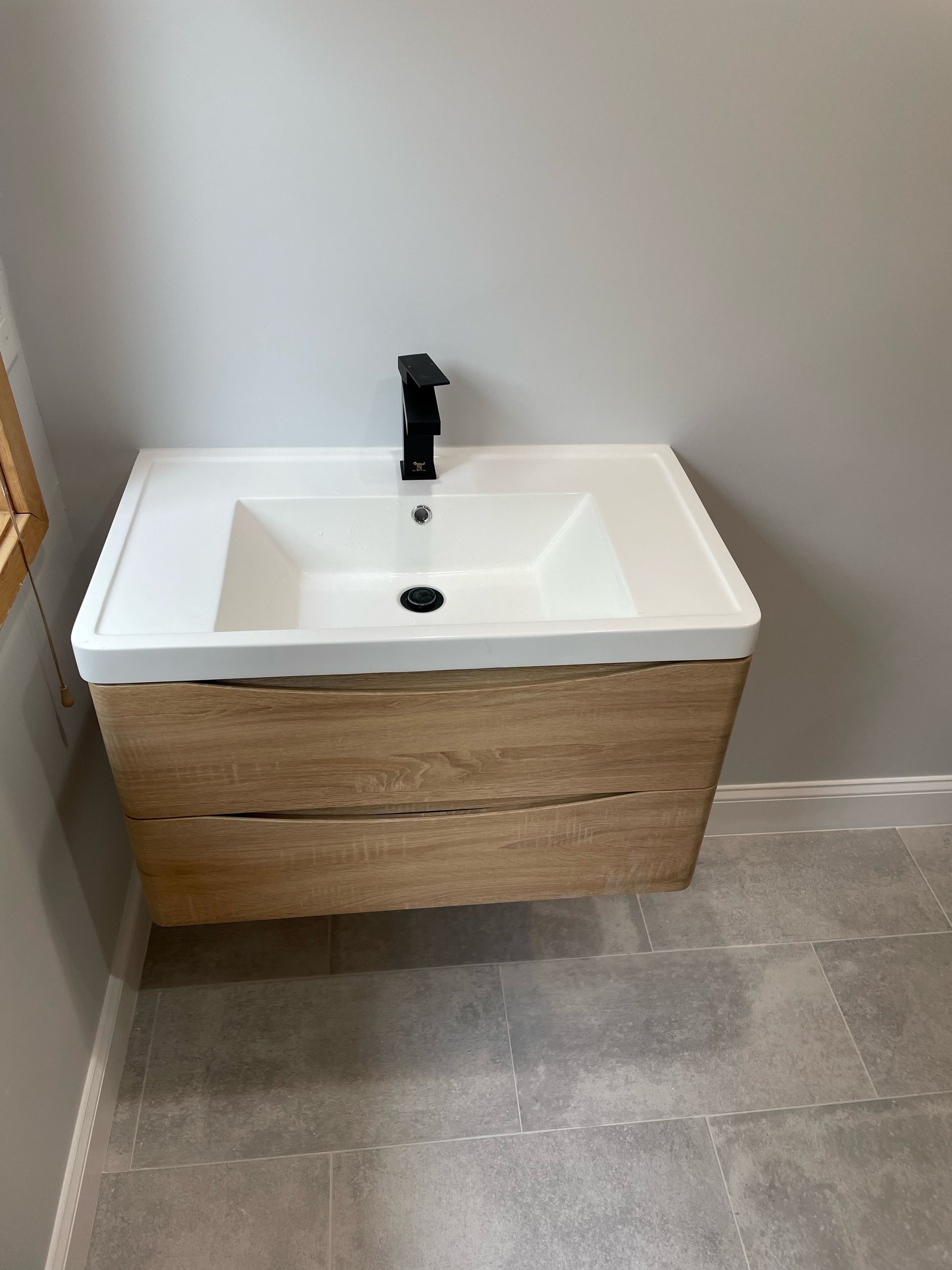 A modern bathroom vanity with wood grain cabinet, white sink, and black faucet on a gray tile floor.