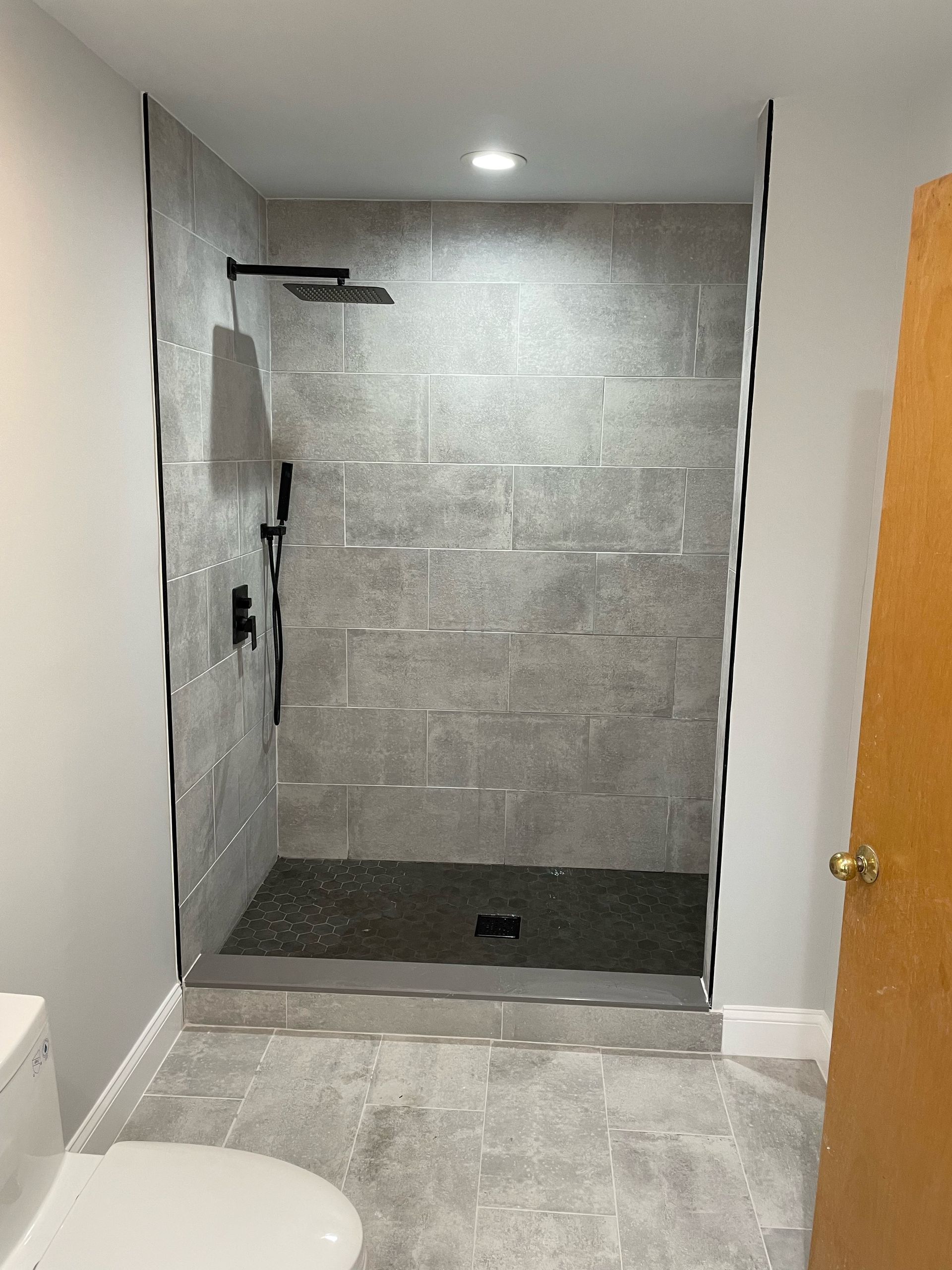 Gray tiled shower stall with black fixtures, open doorway, and toilet.