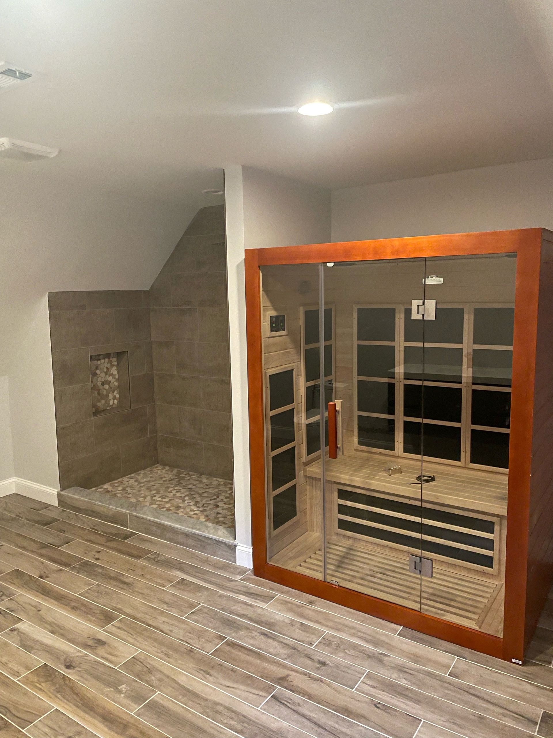 A sauna with a glass door and a shower with stone tiles next to it on a wood floor.