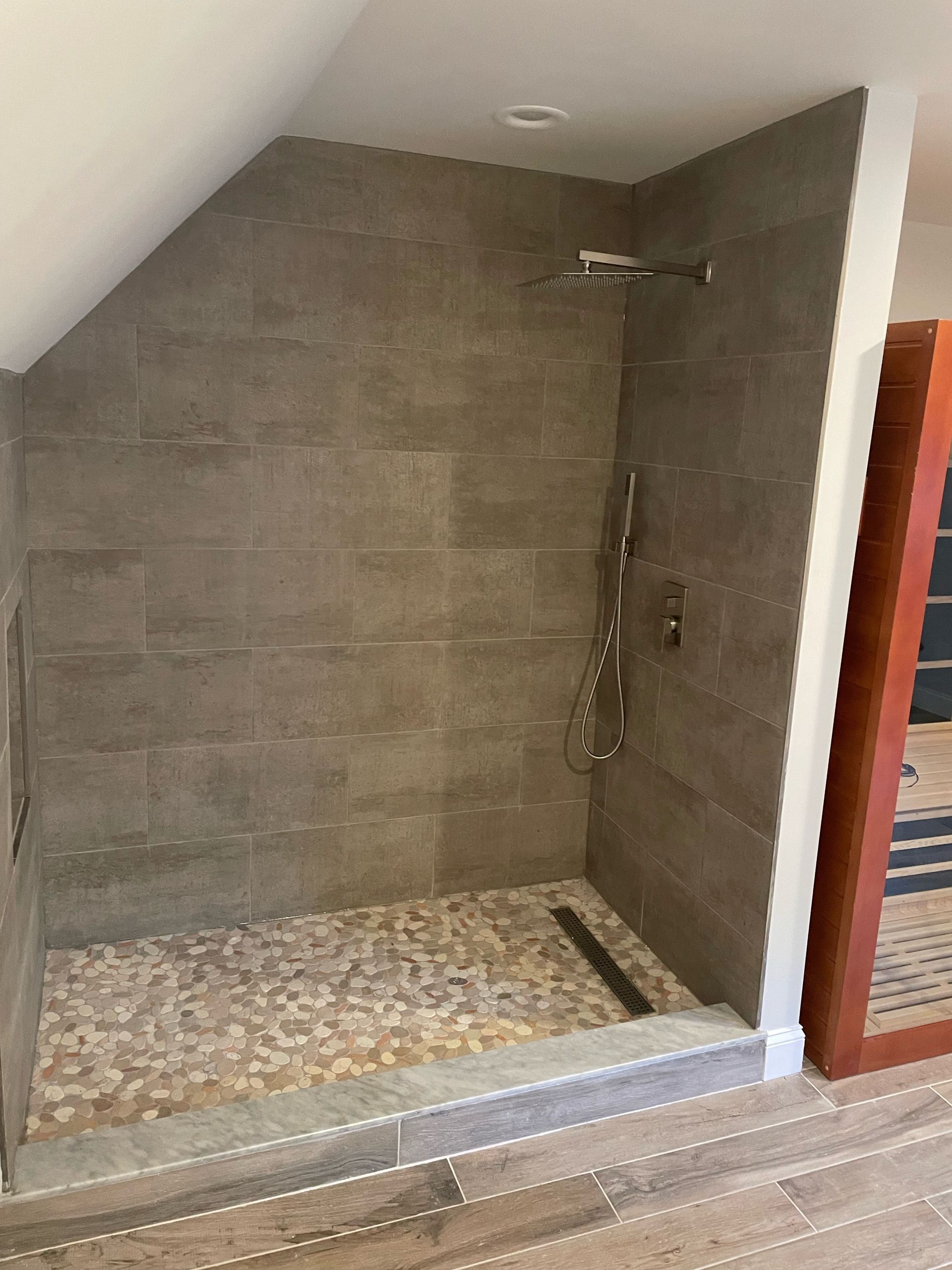 Shower with pebble floor, gray tile walls, rain showerhead, and a sloped ceiling.