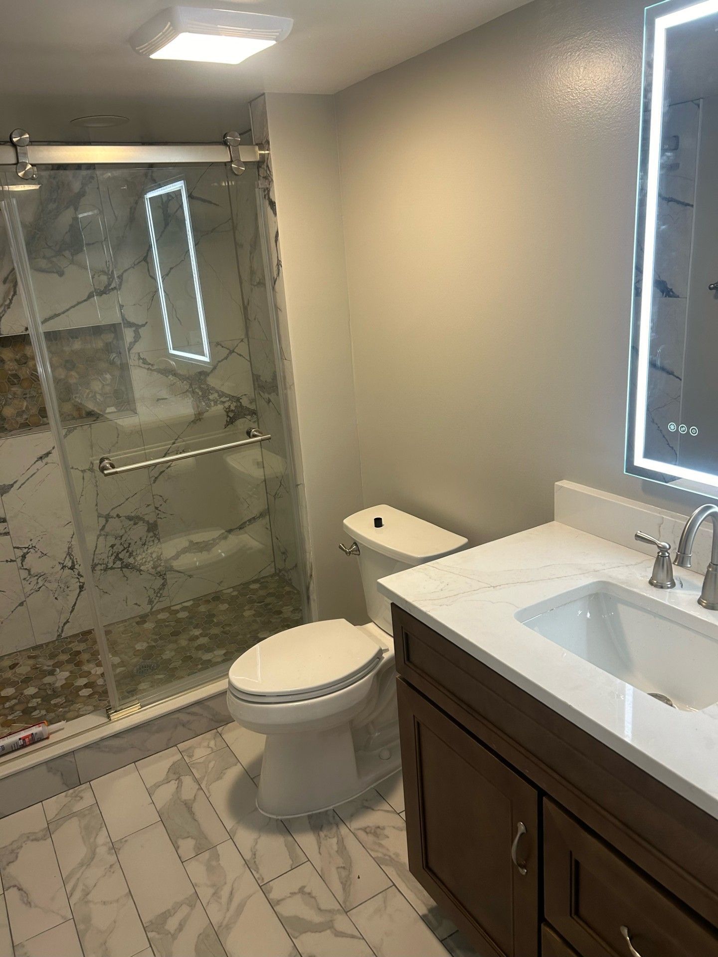 Bathroom with marble-patterned tiles, glass shower, white toilet, wooden vanity, and a lighted mirror.