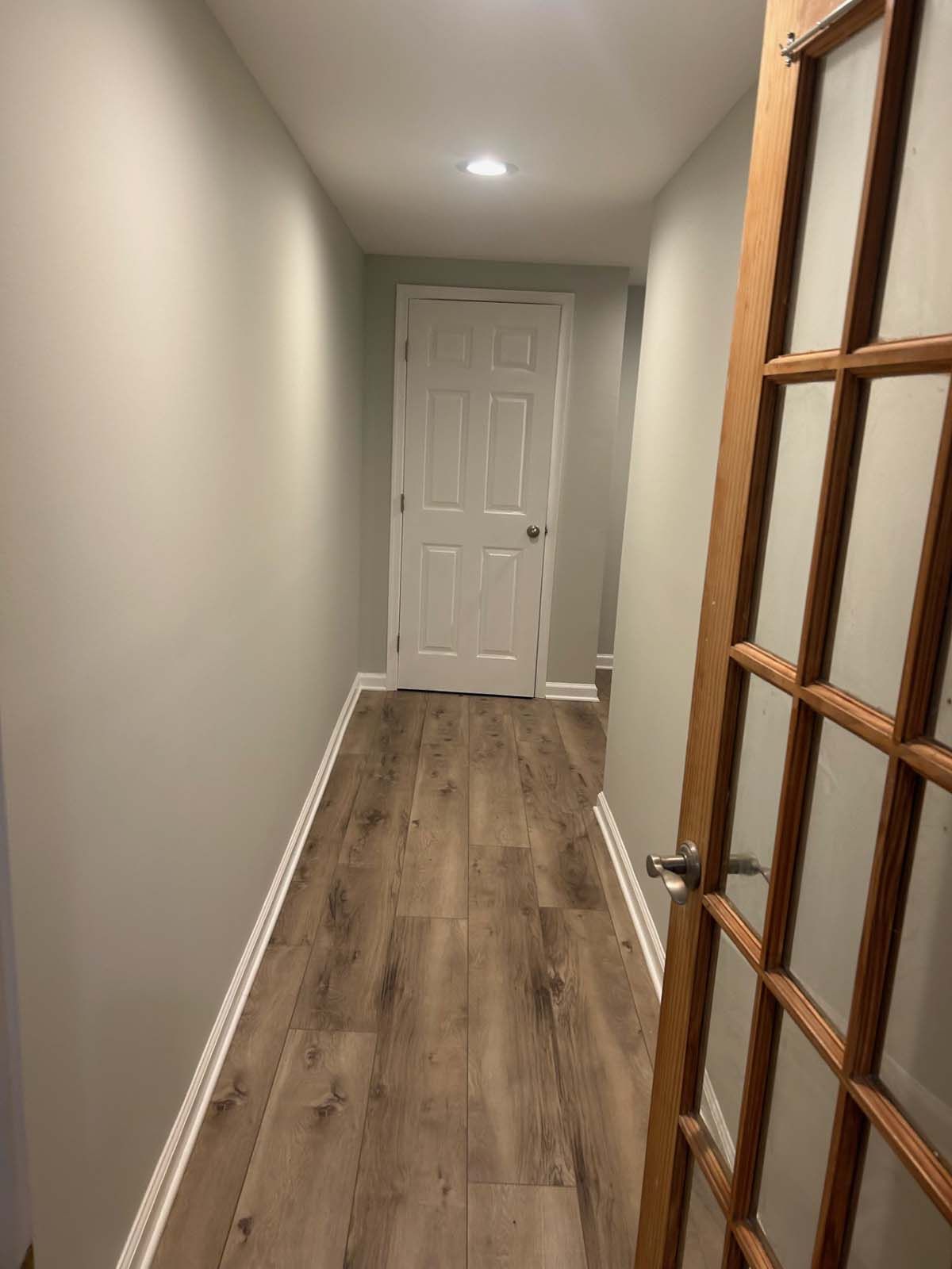 Narrow hallway with wood-look flooring, pale green walls, a white door at the end, and a partially open wood-framed glass door.
