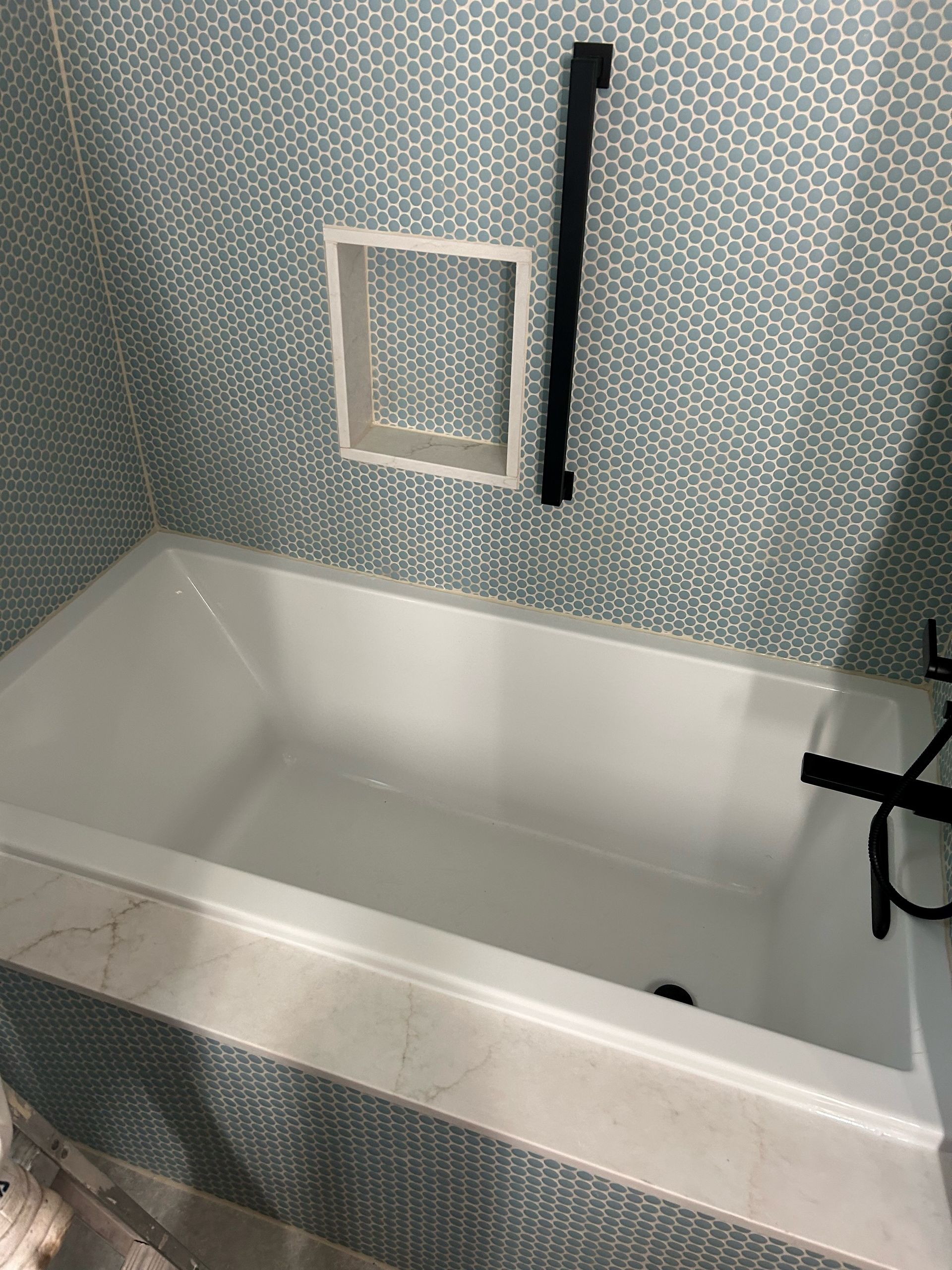 A white bathtub with blue mosaic tile backdrop, a black grab bar, and a recessed shelf.