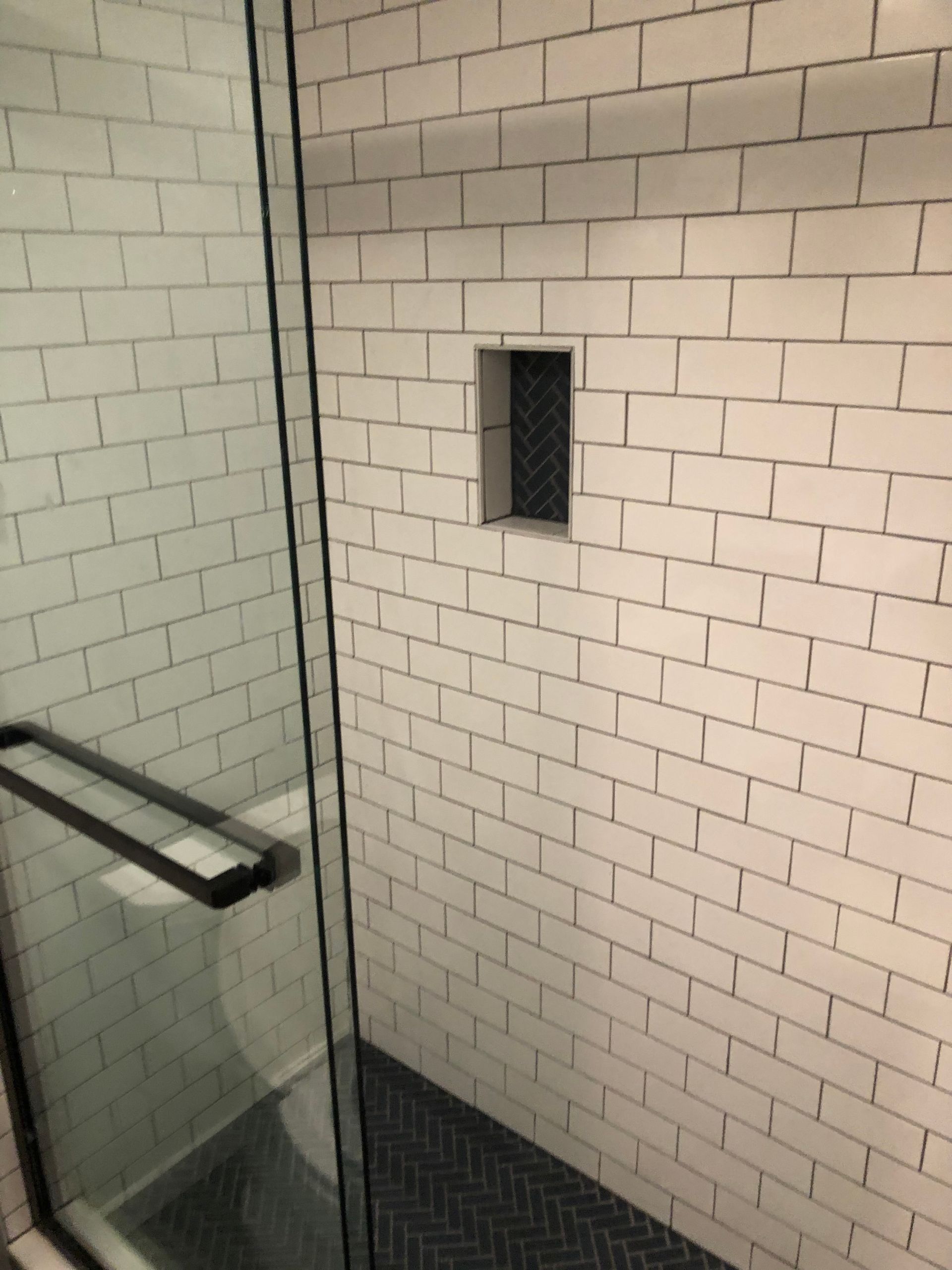 Shower with white brick-like tiles, a black glass door, and a recessed shelf. Black herringbone floor.