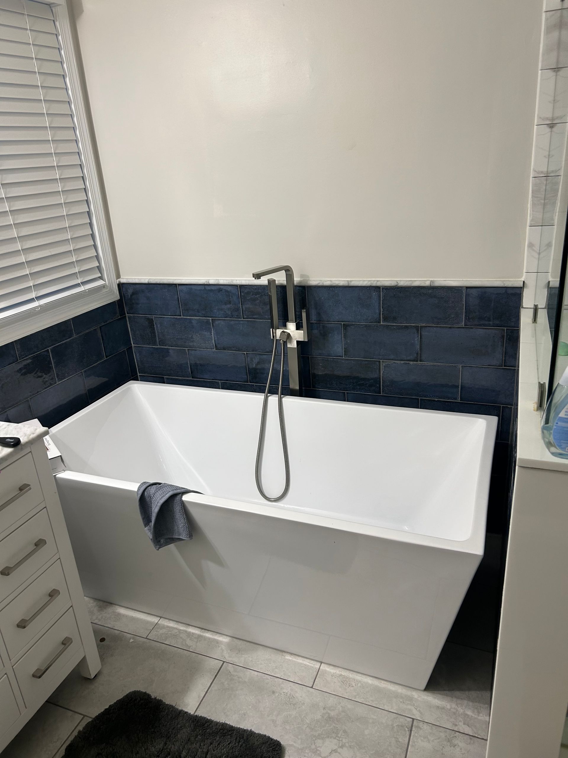 Freestanding white bathtub in a bathroom, navy blue tile backsplash, silver faucet, and towel.