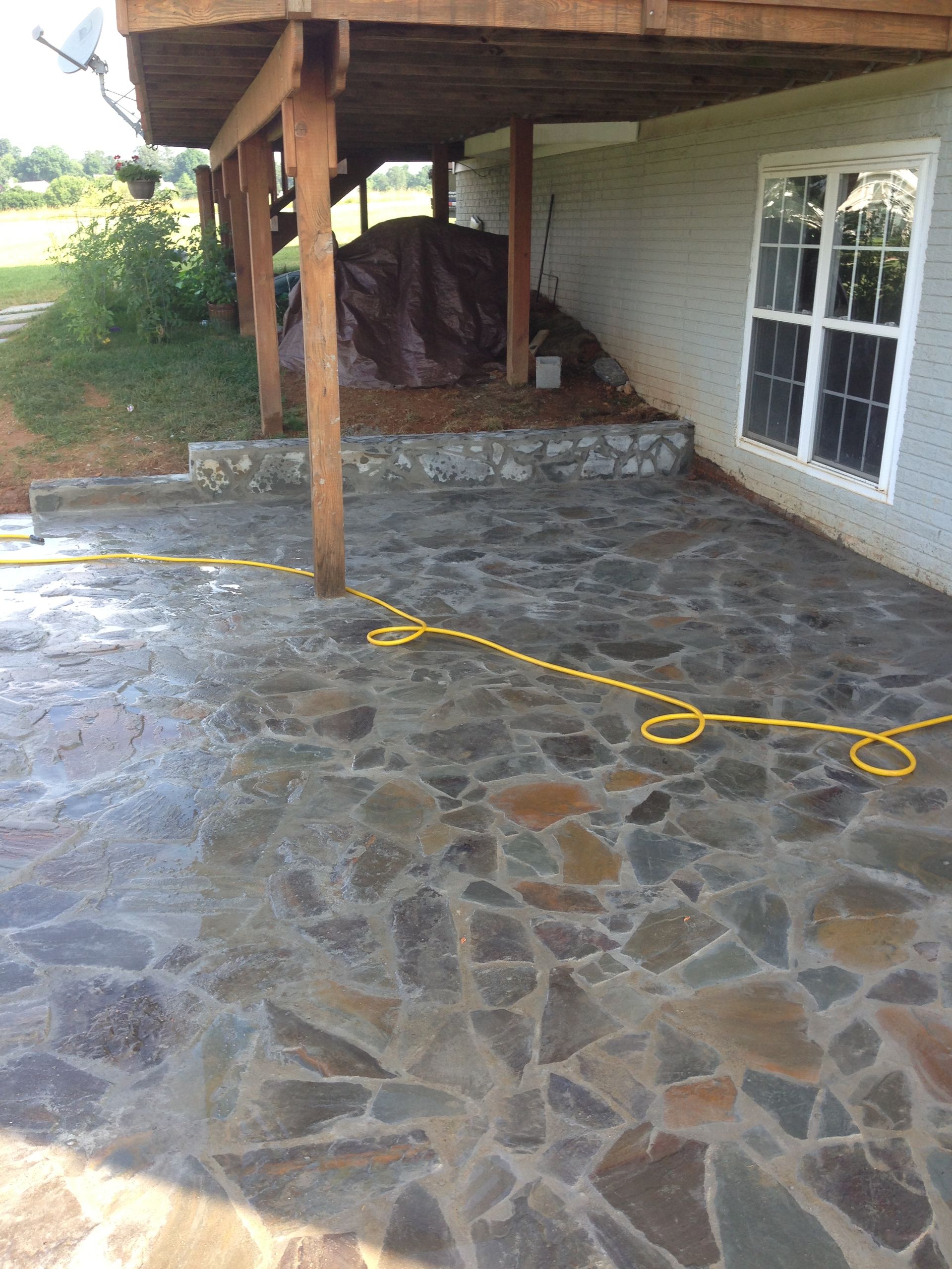 Stone patio under a wooden porch next to a white brick building with a window, a yellow hose lies on the patio.