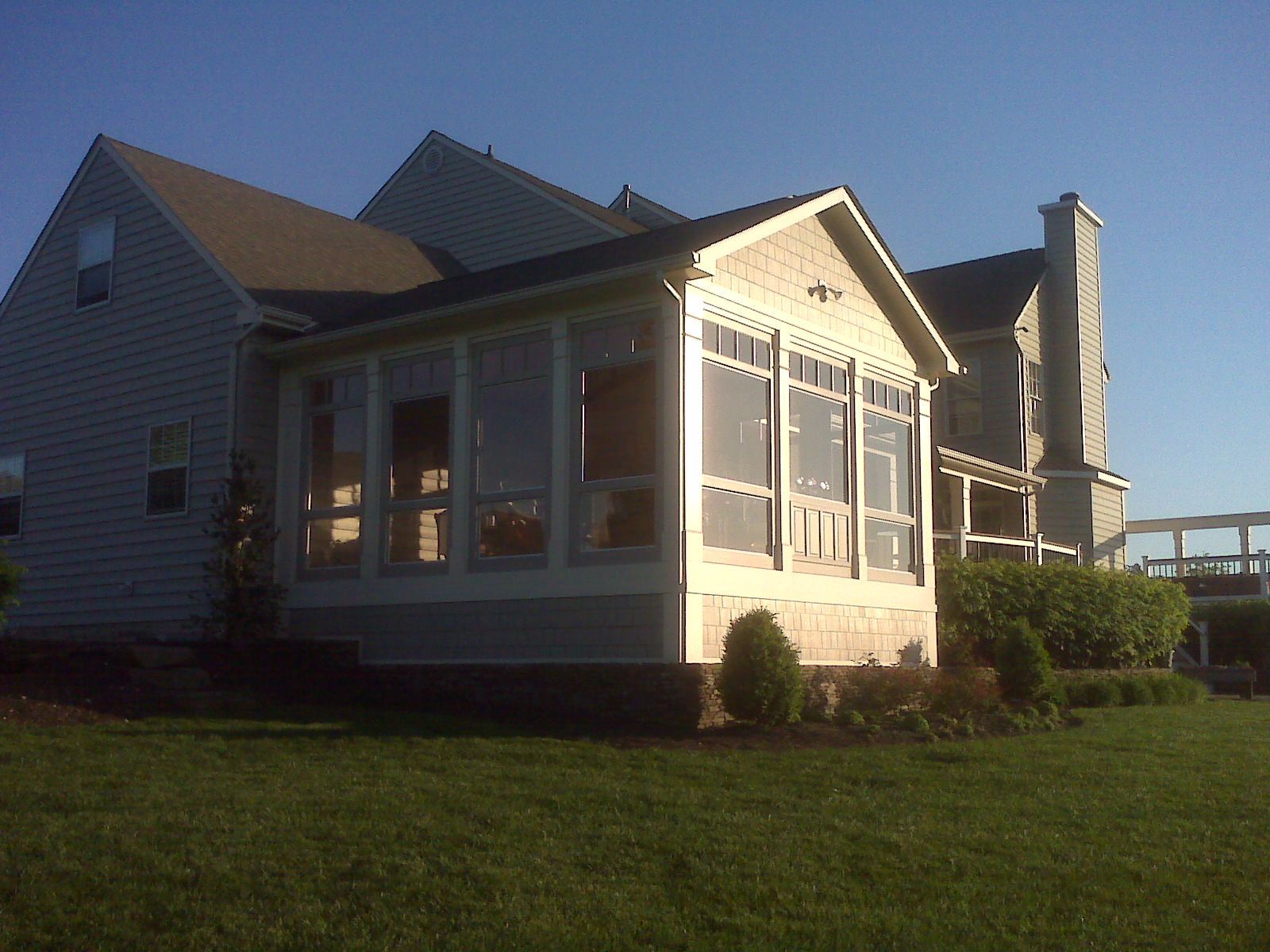 A screened-in porch attached to a light gray house with a chimney, on a green lawn under a blue sky.