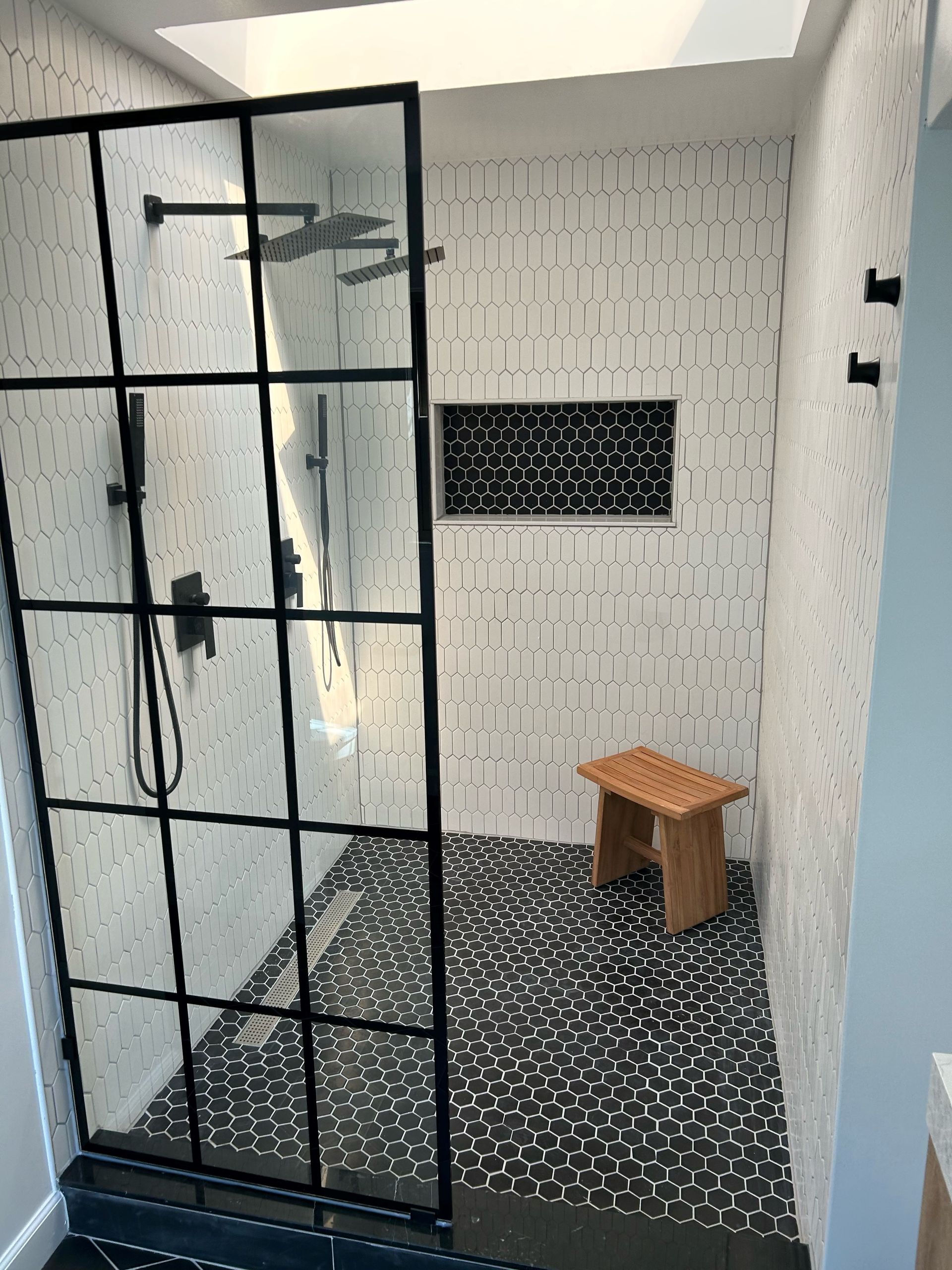 Modern shower with black-framed glass door, patterned floor, white textured tile walls, wooden stool, and black fixtures.