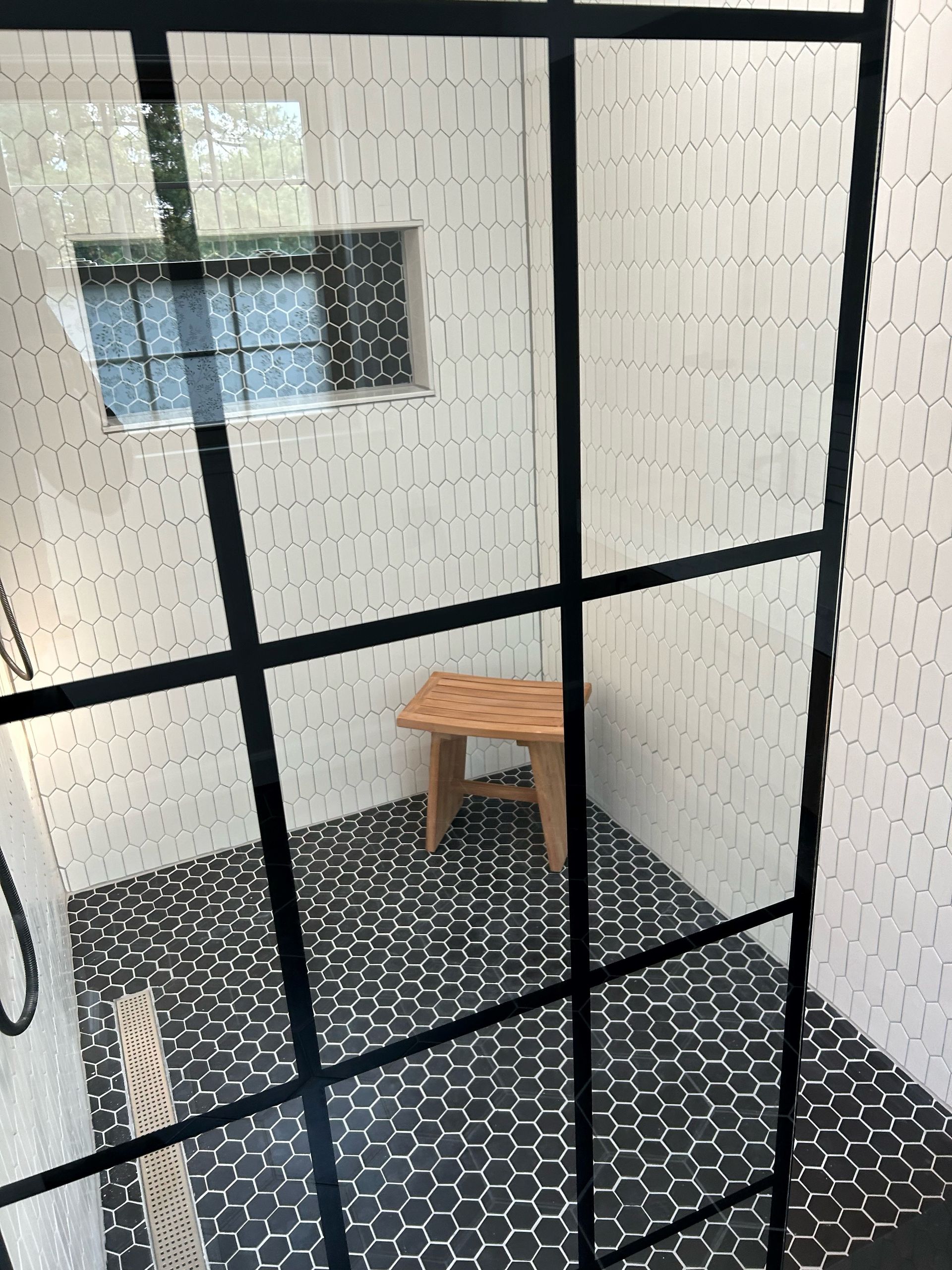 Shower with black and white tile, black metal frame, wooden stool, and small window.
