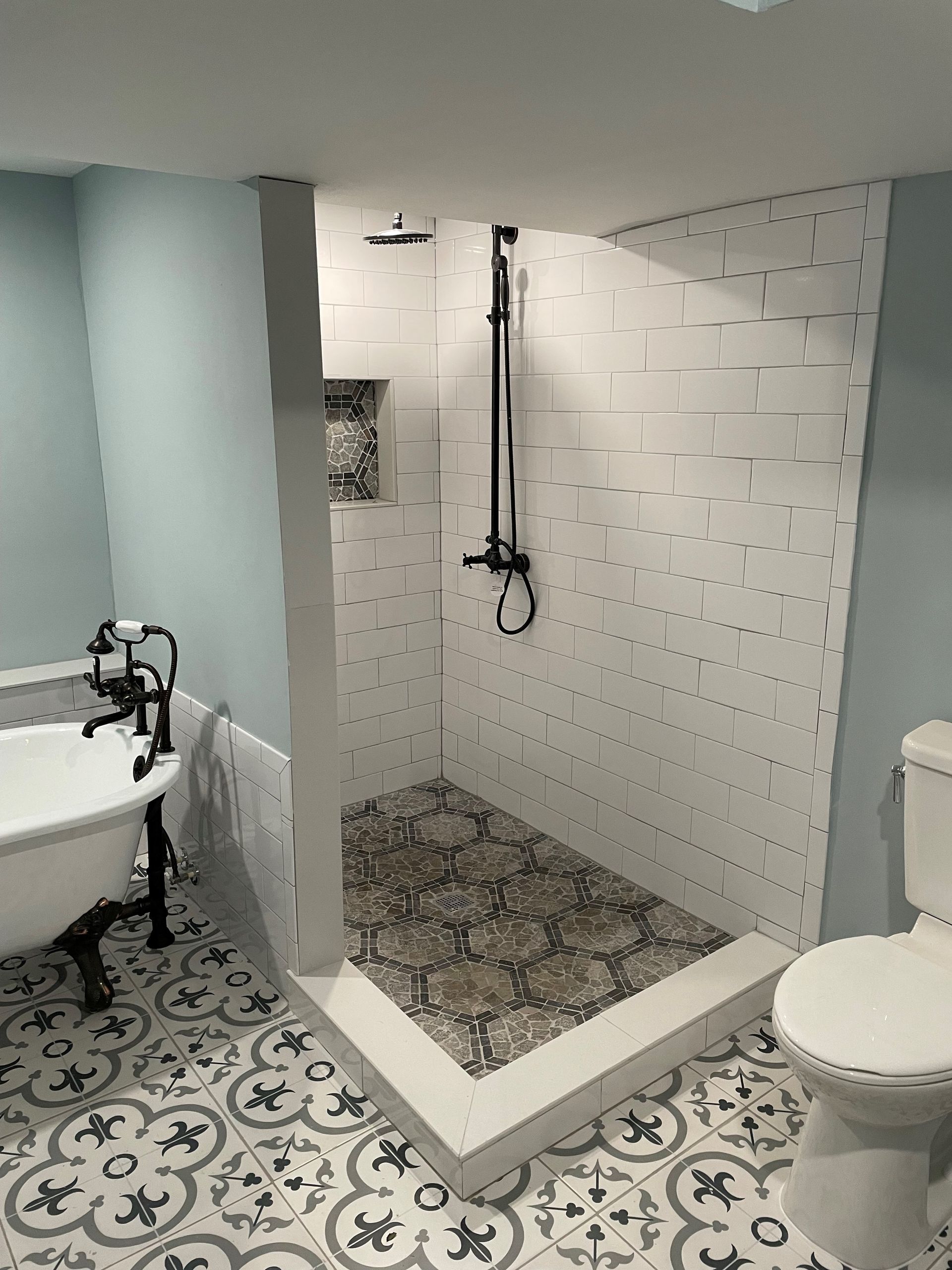 Bathroom with walk-in shower, clawfoot tub, patterned floor tiles, light blue walls, and black fixtures.