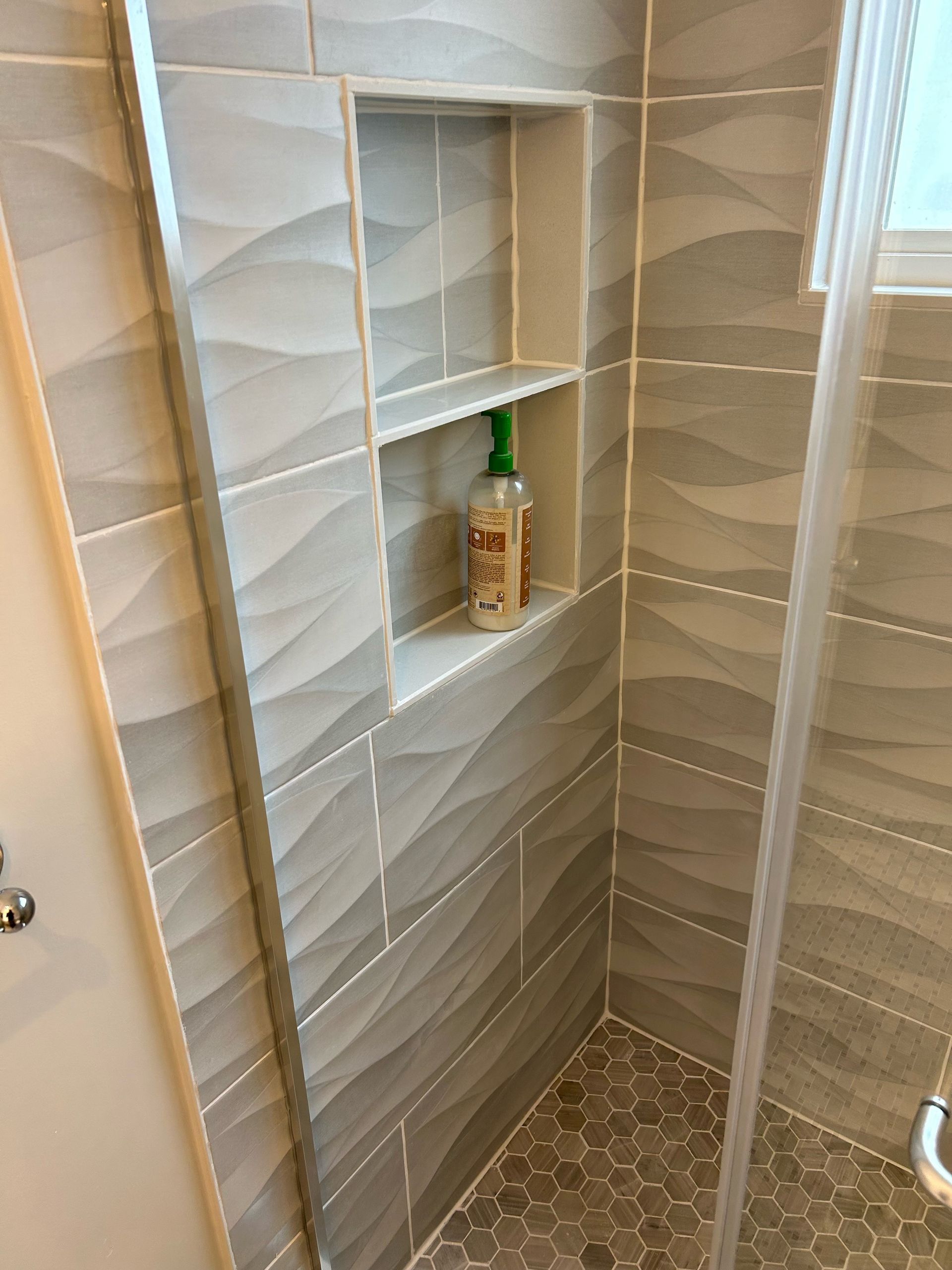 Gray tiled shower with a built-in shelf holding a shampoo bottle.