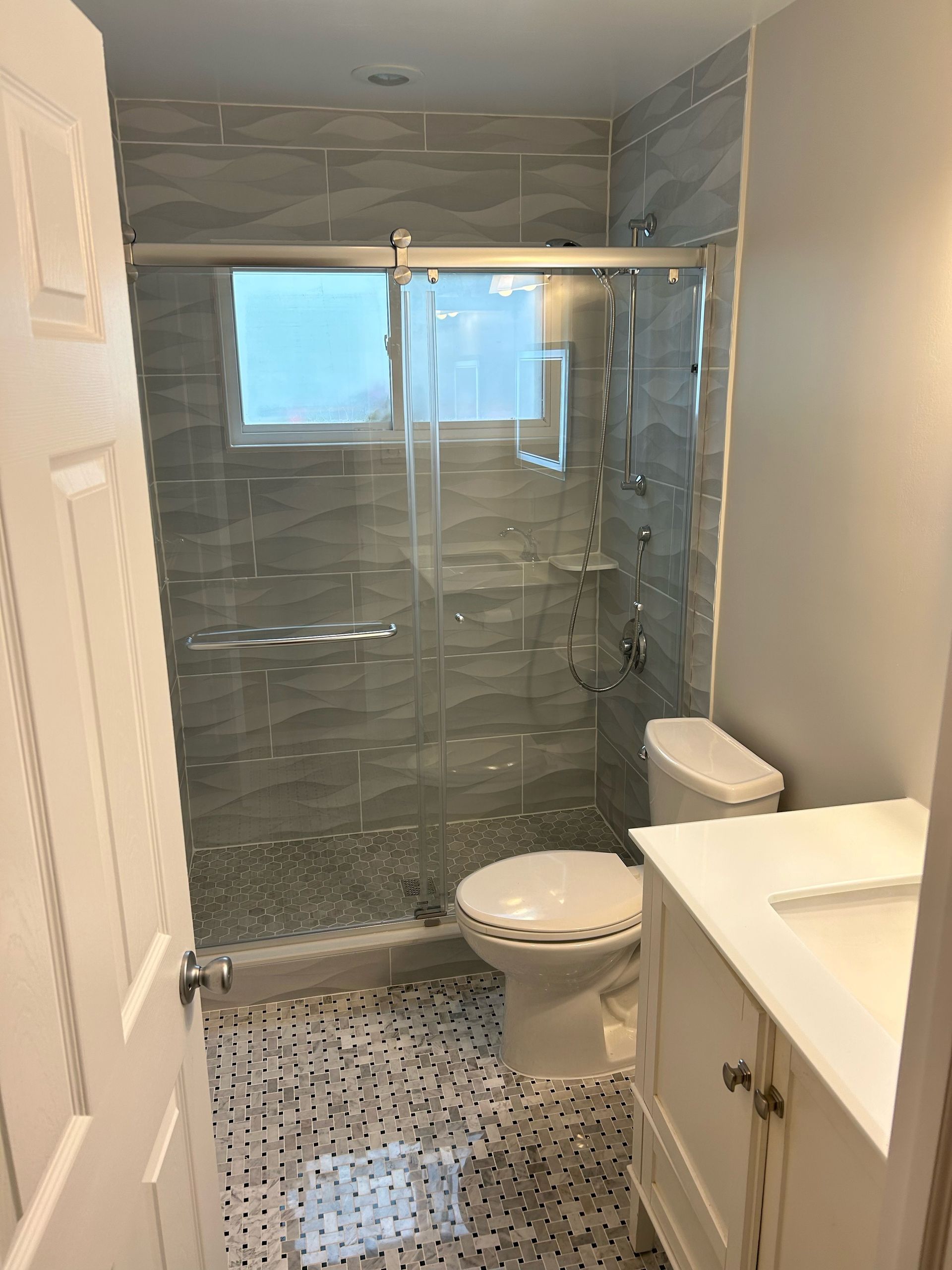 Bathroom with gray tiled walls, glass shower, white toilet and vanity, patterned floor.