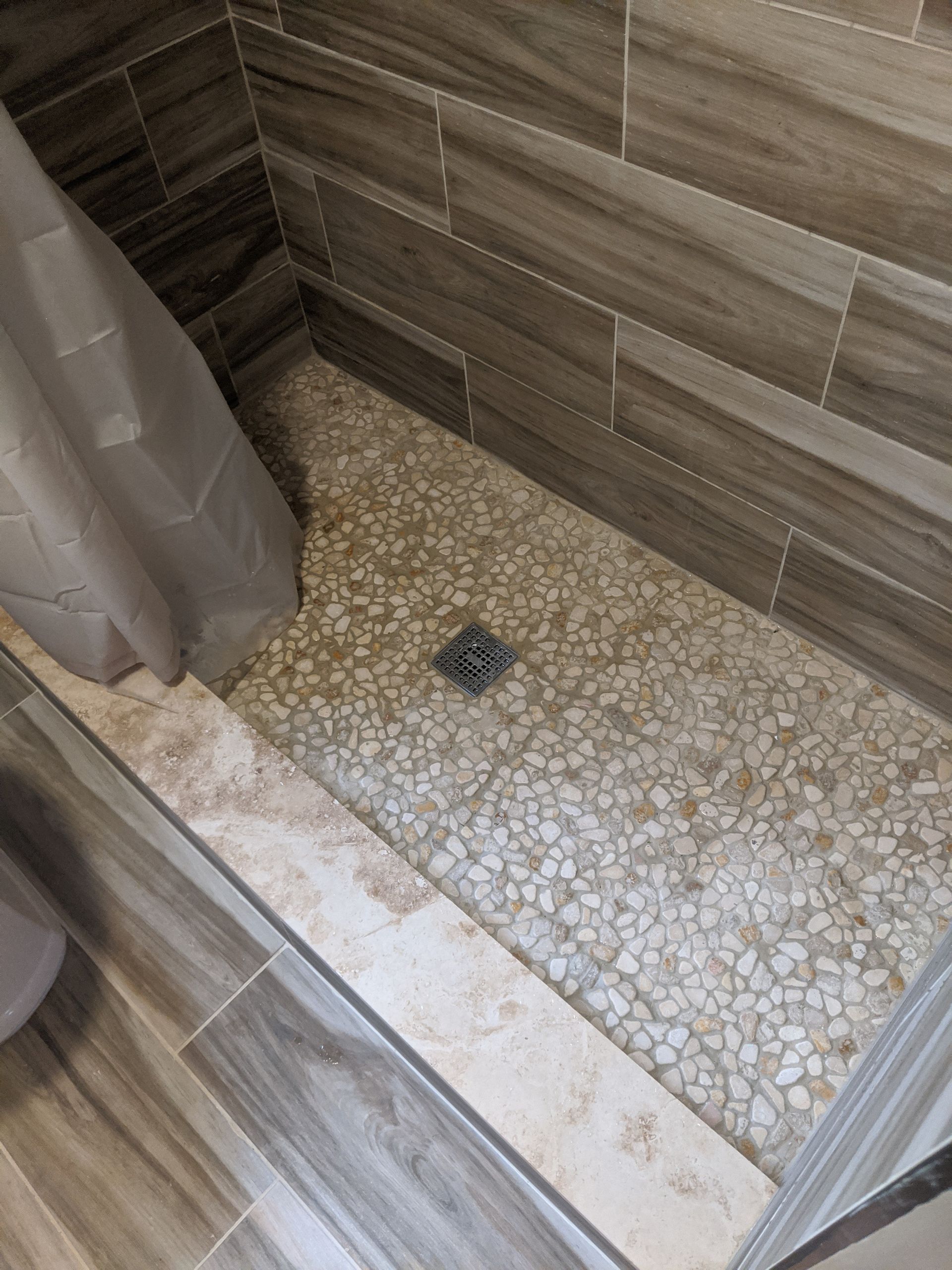 Shower stall with pebble tile floor and wood-look wall tiles, curtain to the left, and a drain in the center.