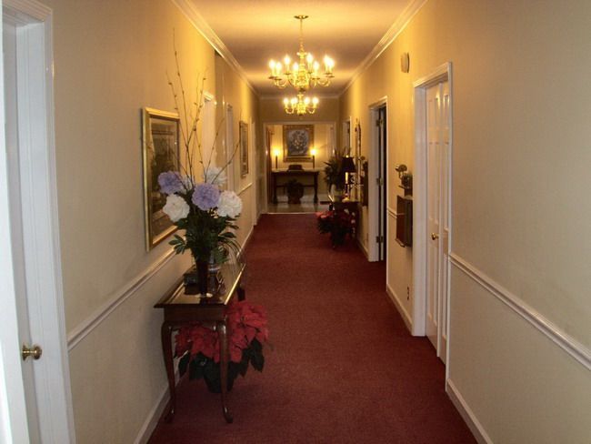 Long hallway with red carpet, cream walls, framed artwork, and chandelier.