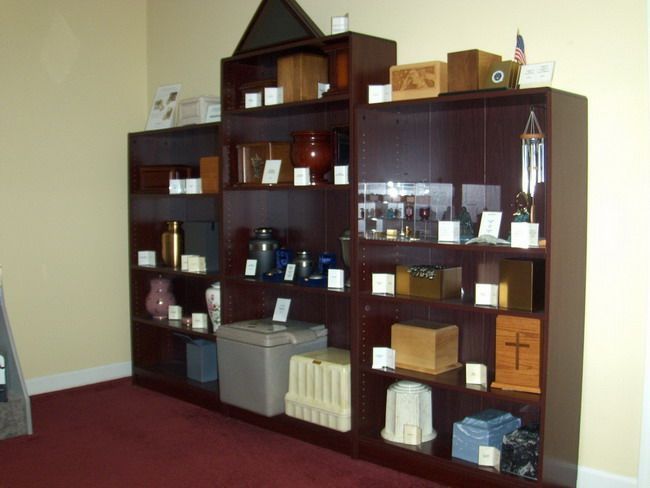 Dark wooden shelves displaying various cremation urns and keepsakes in a room with a red carpet.