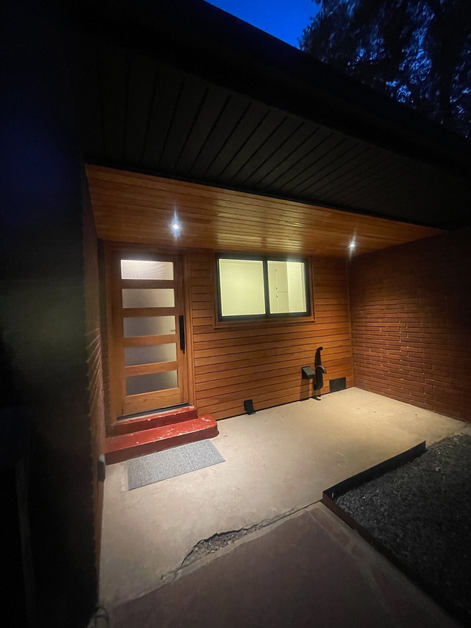 Lit porch with wooden door, window, and red brick wall; at night.