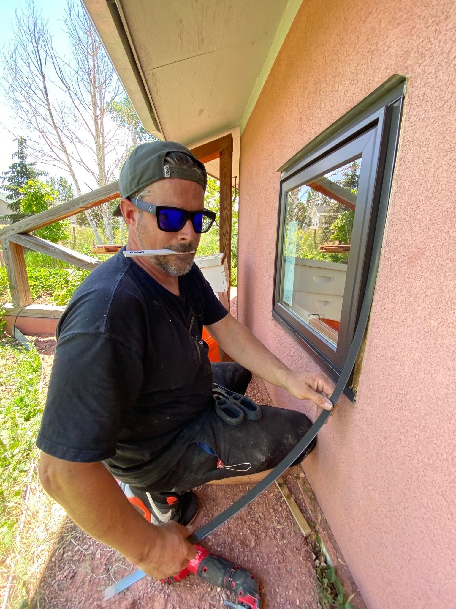 Man installing black window trim on a pink house exterior, using a saw.