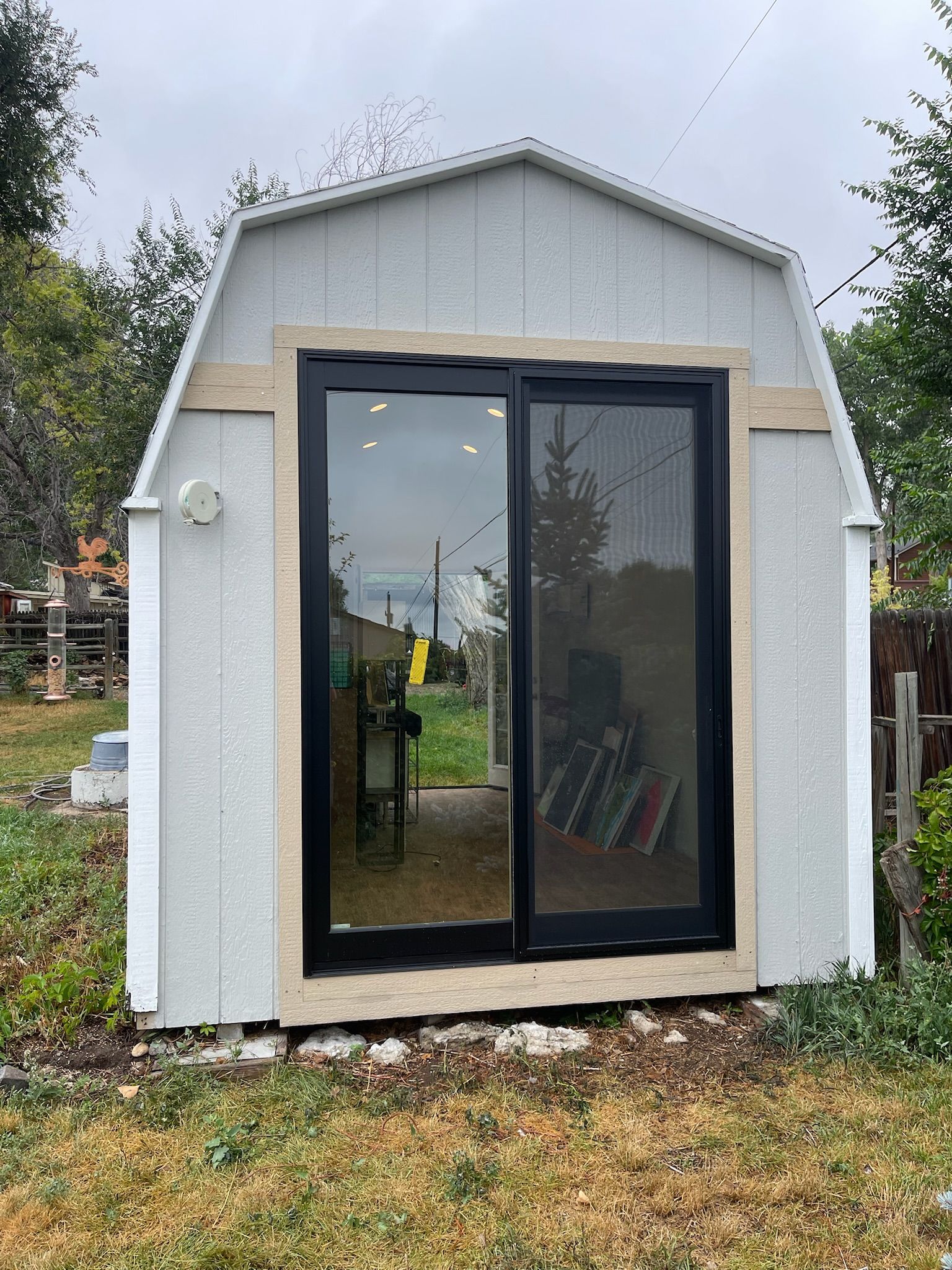 Gray shed with a black-framed sliding glass door. Light wood trim surrounds the door. Overcast sky.
