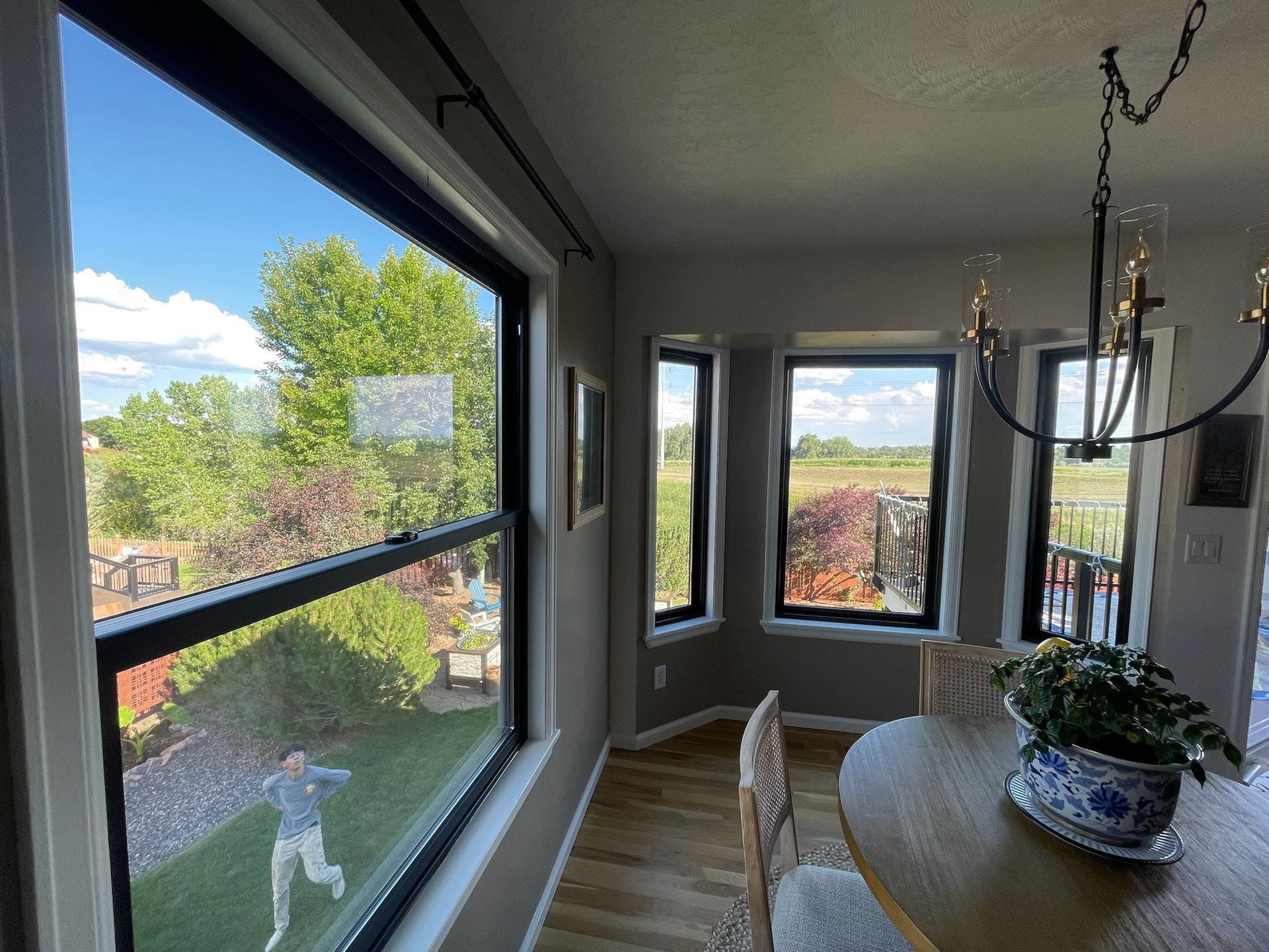Interior view of a dining area with large windows overlooking a yard and trees. A person appears to be playing in the yard.