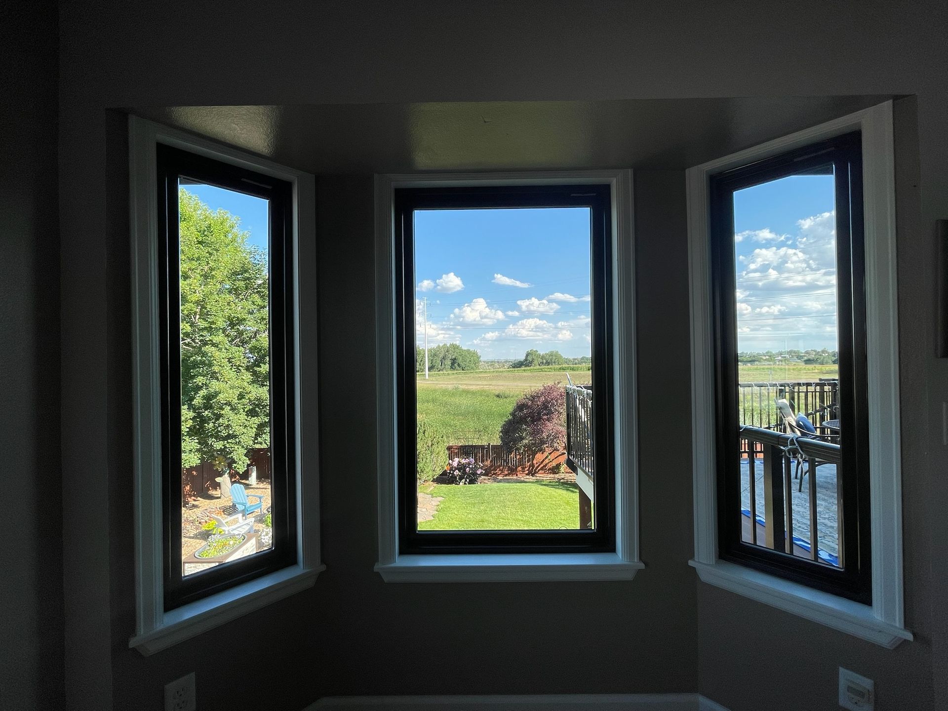 Three windows with black frames showing a bright outdoor view of trees, grass, and a waterway under a blue sky.